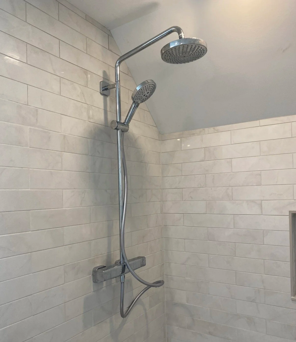 Bathroom shower with a dual showerhead system mounted on a tiled wall, featuring a large rain showerhead and a smaller handheld showerhead.