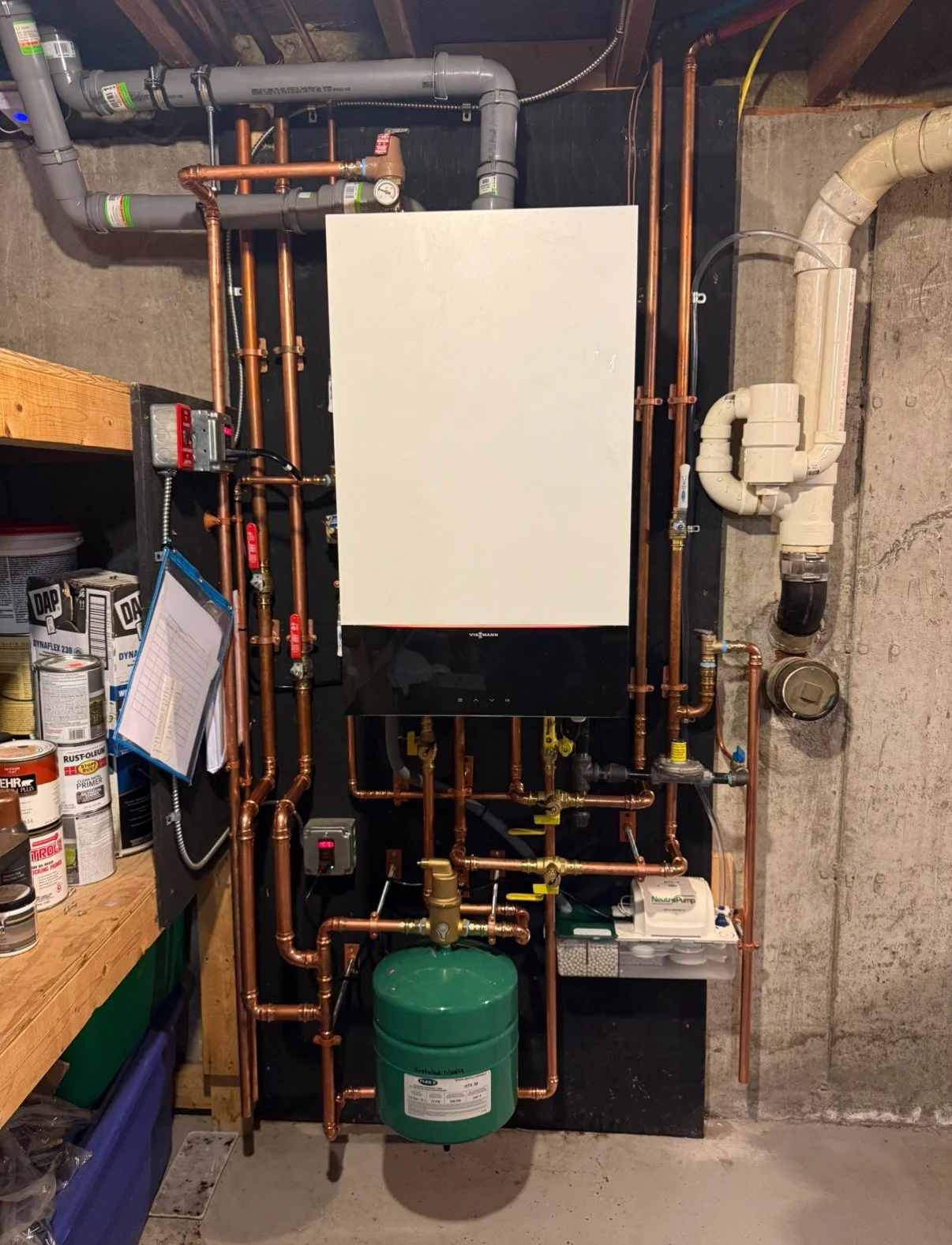 This is a mechanical room with copper piping, a white boiler, and a green expansion tank, all mounted on a concrete wall.