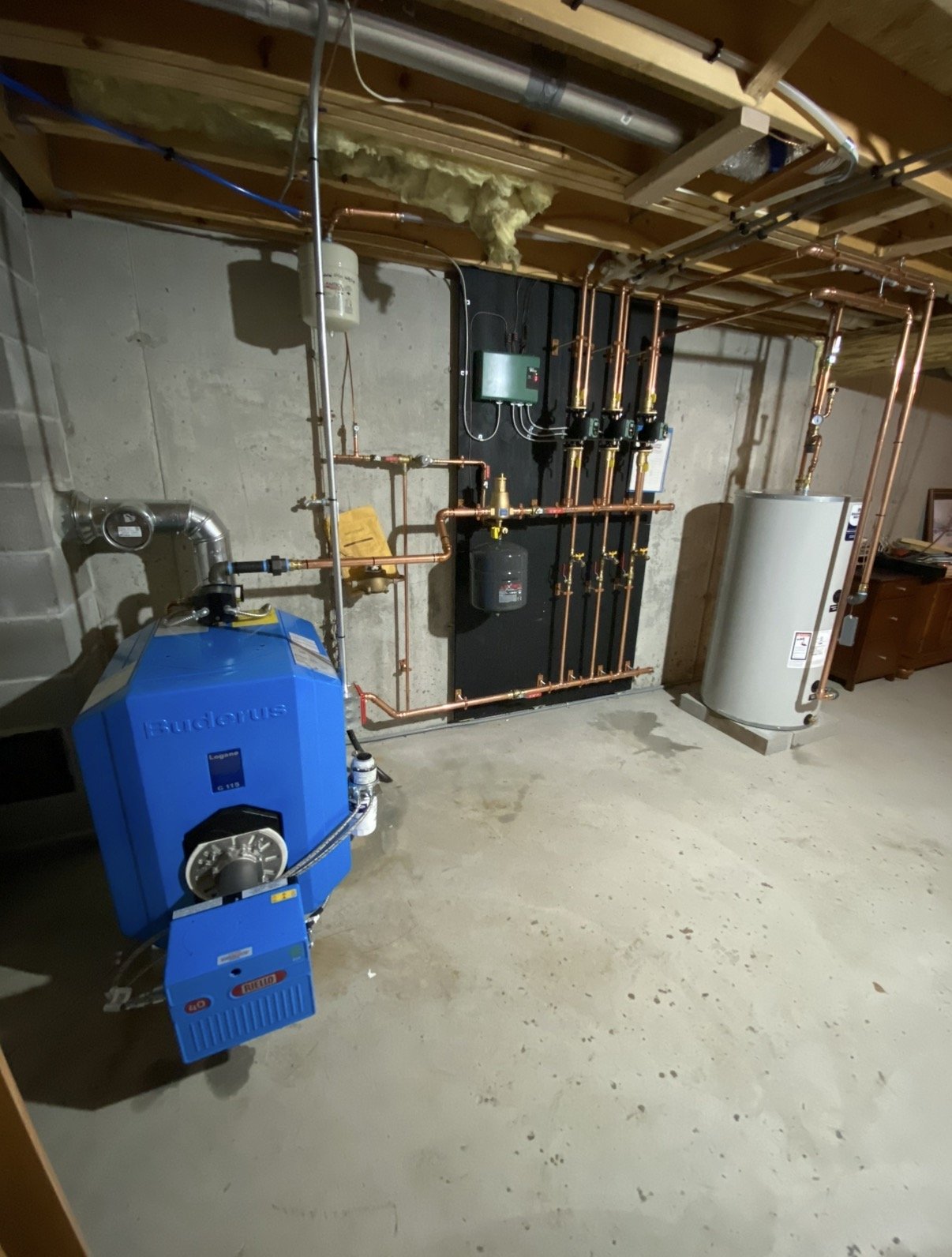 Basement with a water heater, copper piping, and a blue boiler system installed on a concrete floor and unfinished ceiling.