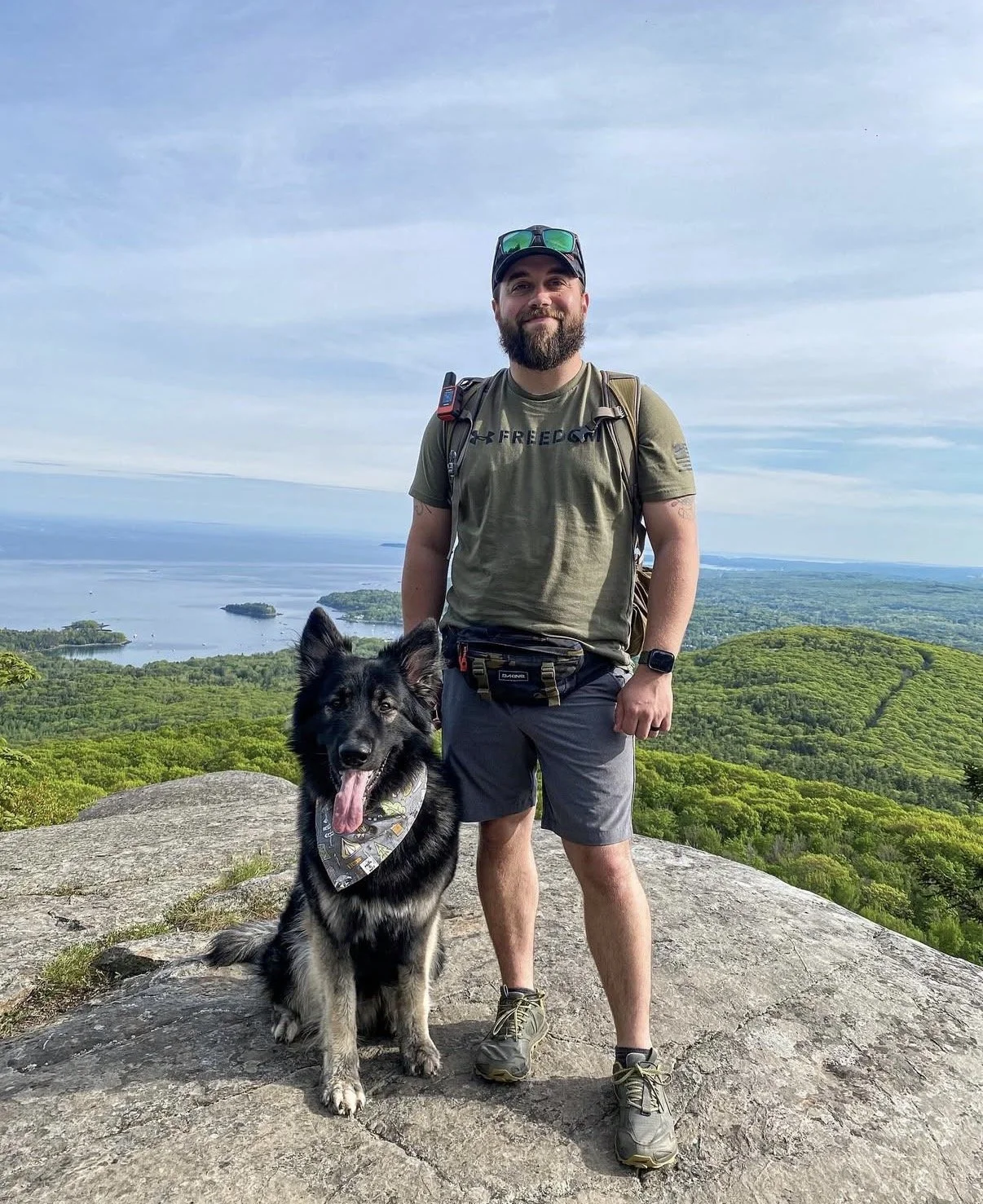 A man with a beard and a dog standing on a rocky hilltop with a scenic view of a lake, small islands, and green forests in the background.