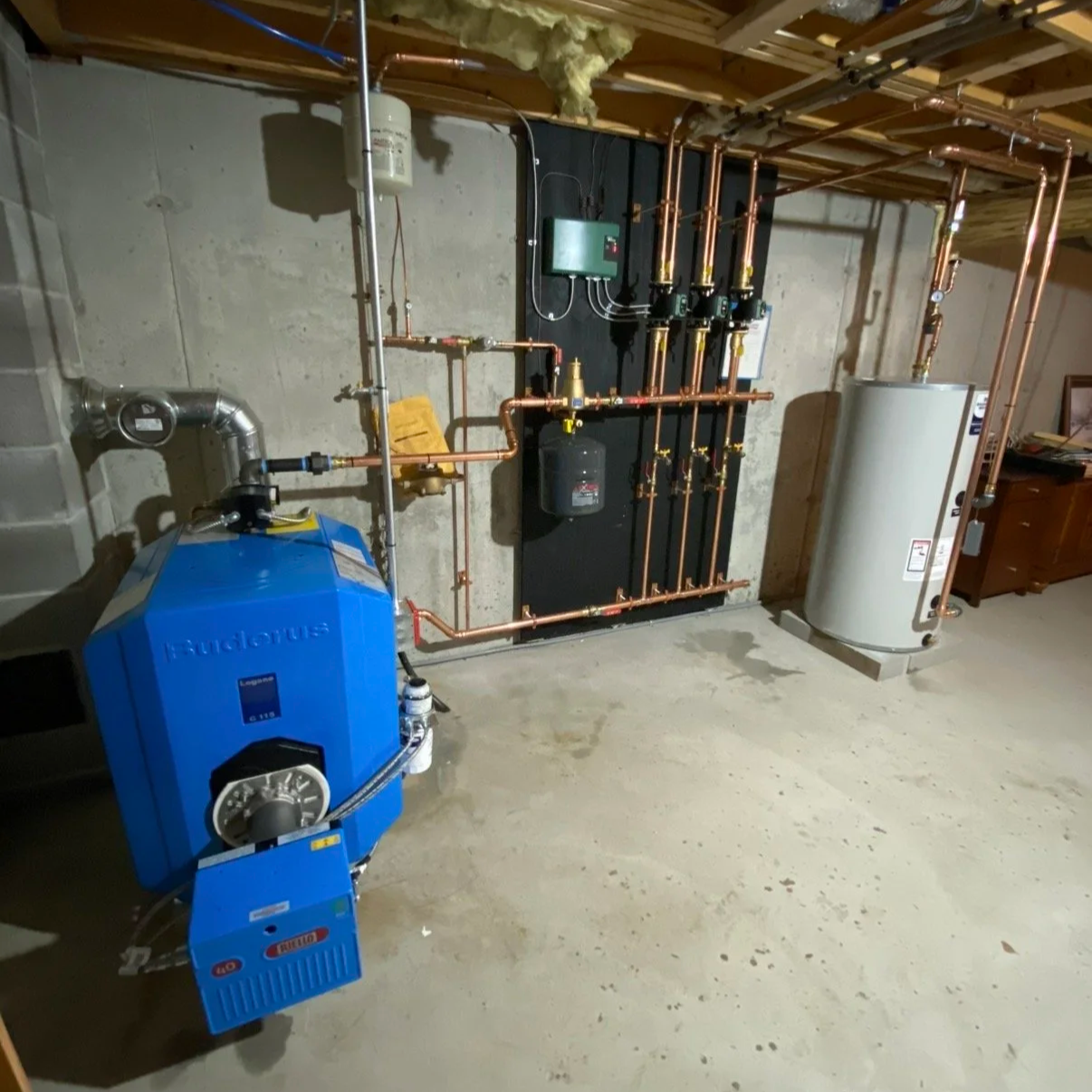 A basement room with a Lennox blue boiler, a white water heater, and copper piping connected to other mechanical equipment, including a black control panel mounted on a black wall panel.