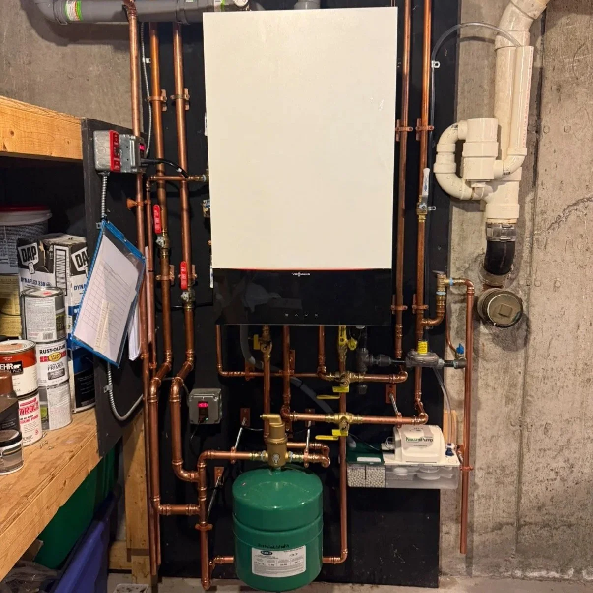 An indoor plumbing and heating system with copper pipes, a green expansion tank, various valves, and a digital control panel mounted on a wall next to a concrete wall and shelves with cans of paint and other supplies.