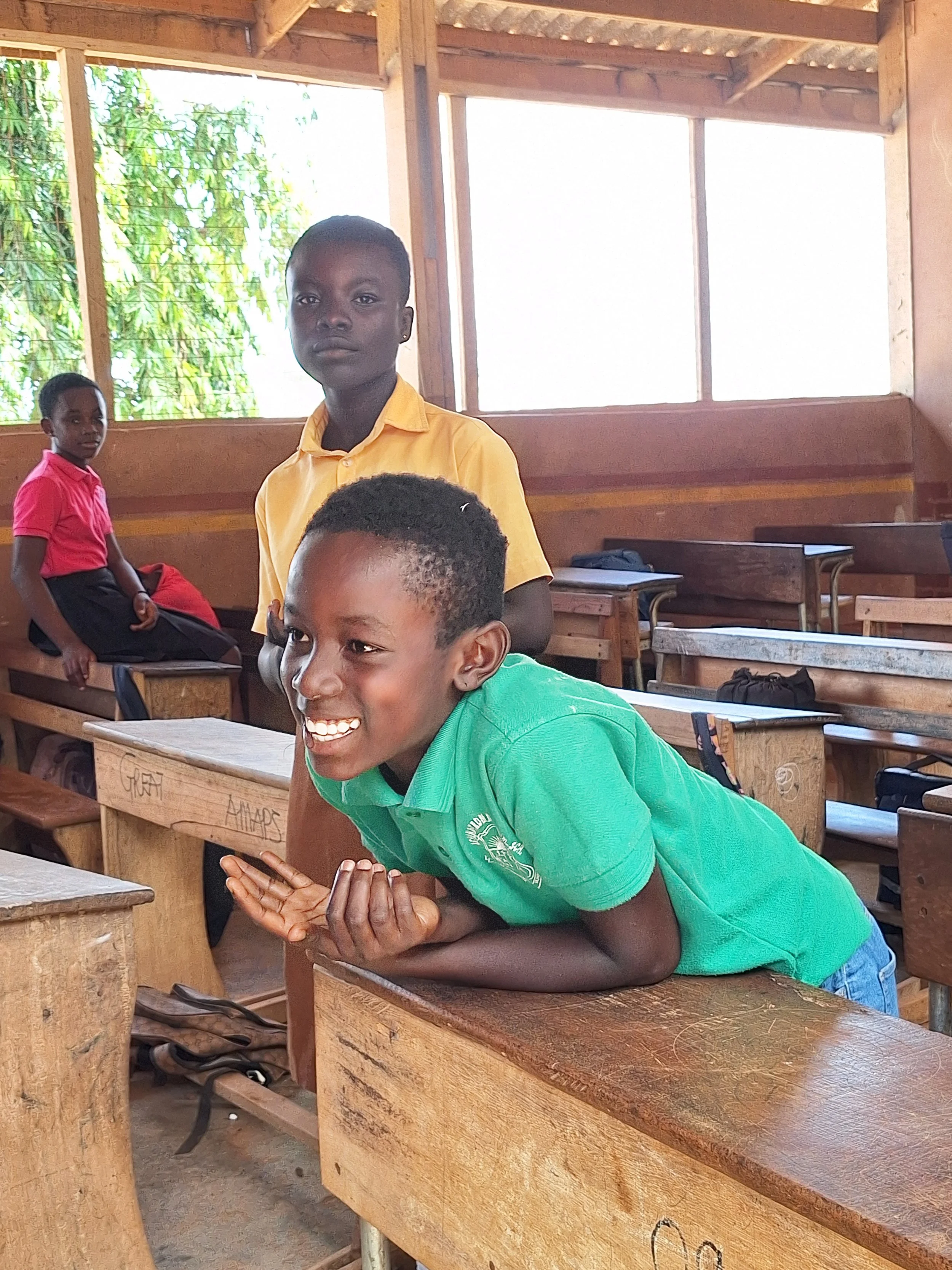 Three students inside a wooden classroom, one in a green shirt leaning on a desk smiling, one in a yellow shirt standing looking at the camera, and another girl in a pink shirt sitting on a desk in the background.