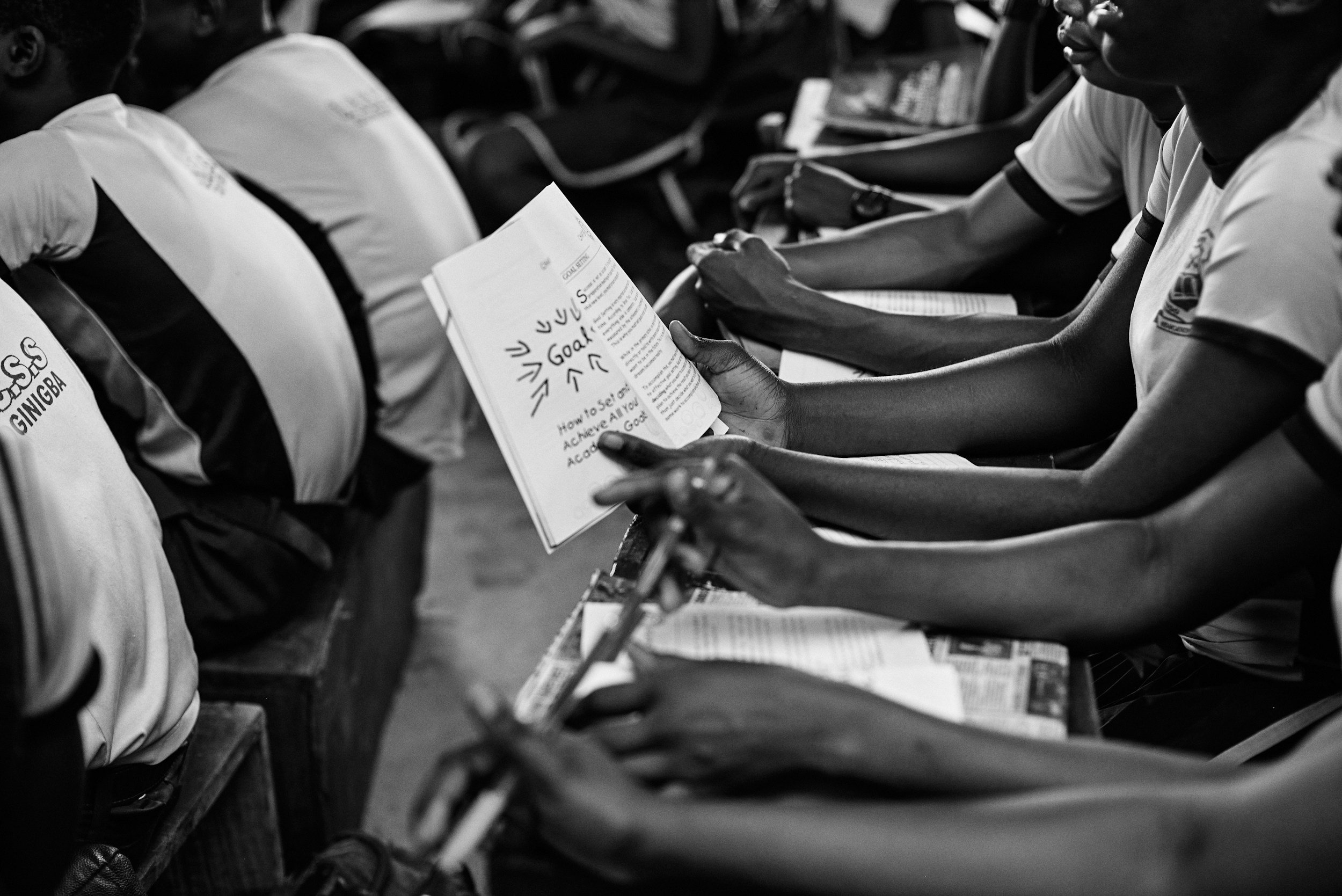 People seated in a row, reading booklets, in a black-and-white photo.