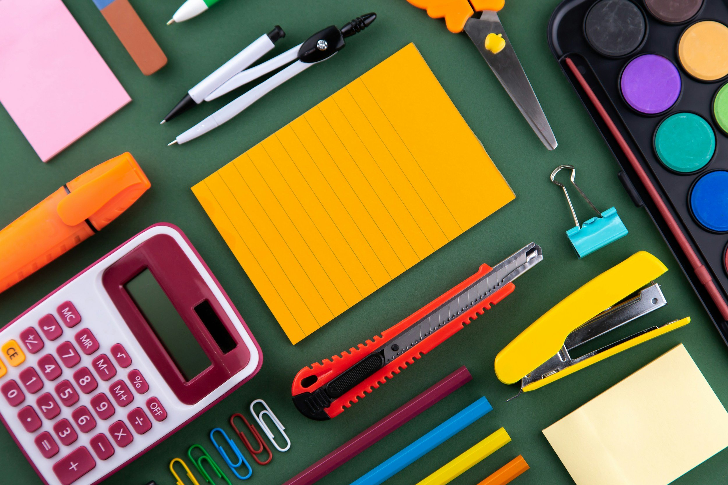 Colorful office or school supplies including notebooks, pens, a calculator, scissors, paper clips, markers, a water color palette, and a stapler arranged on a green background.