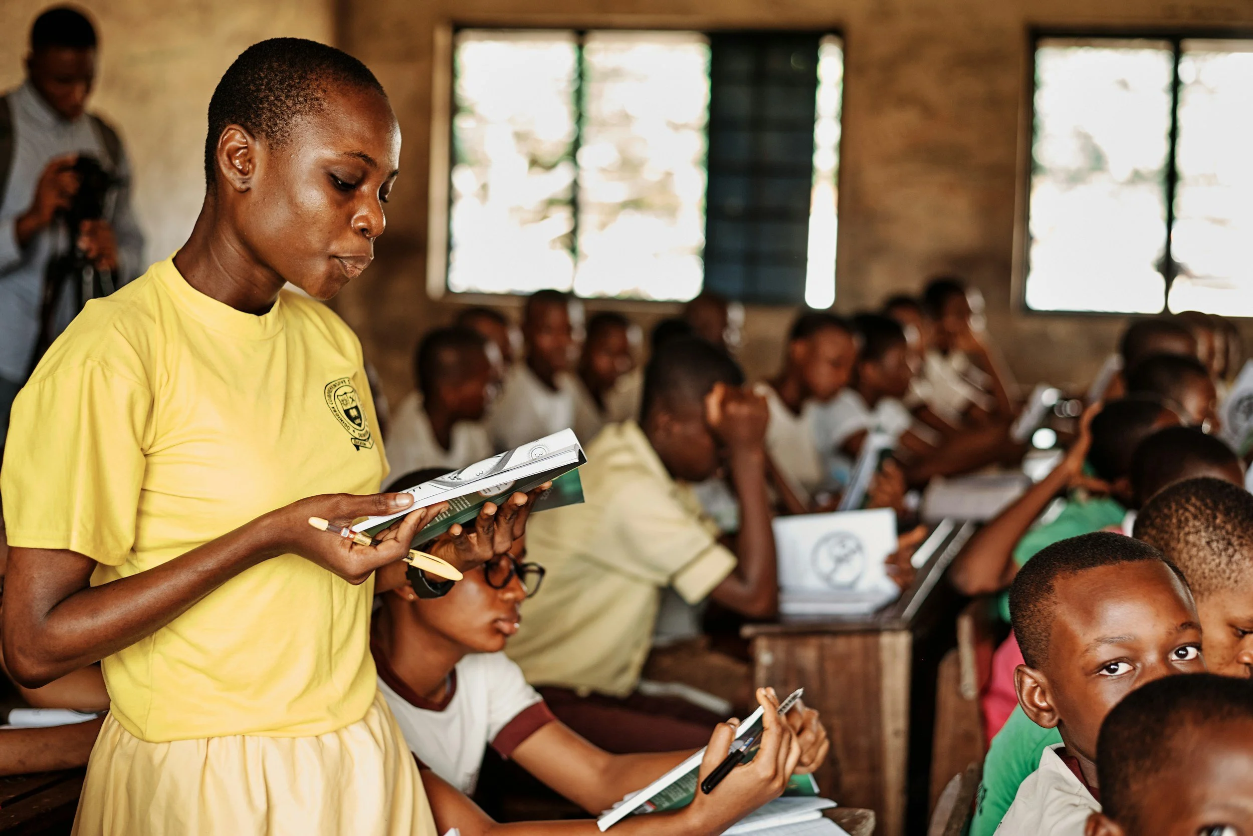 A woman in a yellow shirt standing in a classroom filled with students. She is reading from a book or notebook. Other students are seated, paying attention, and some are holding notebooks or tablets. The classroom has large windows.