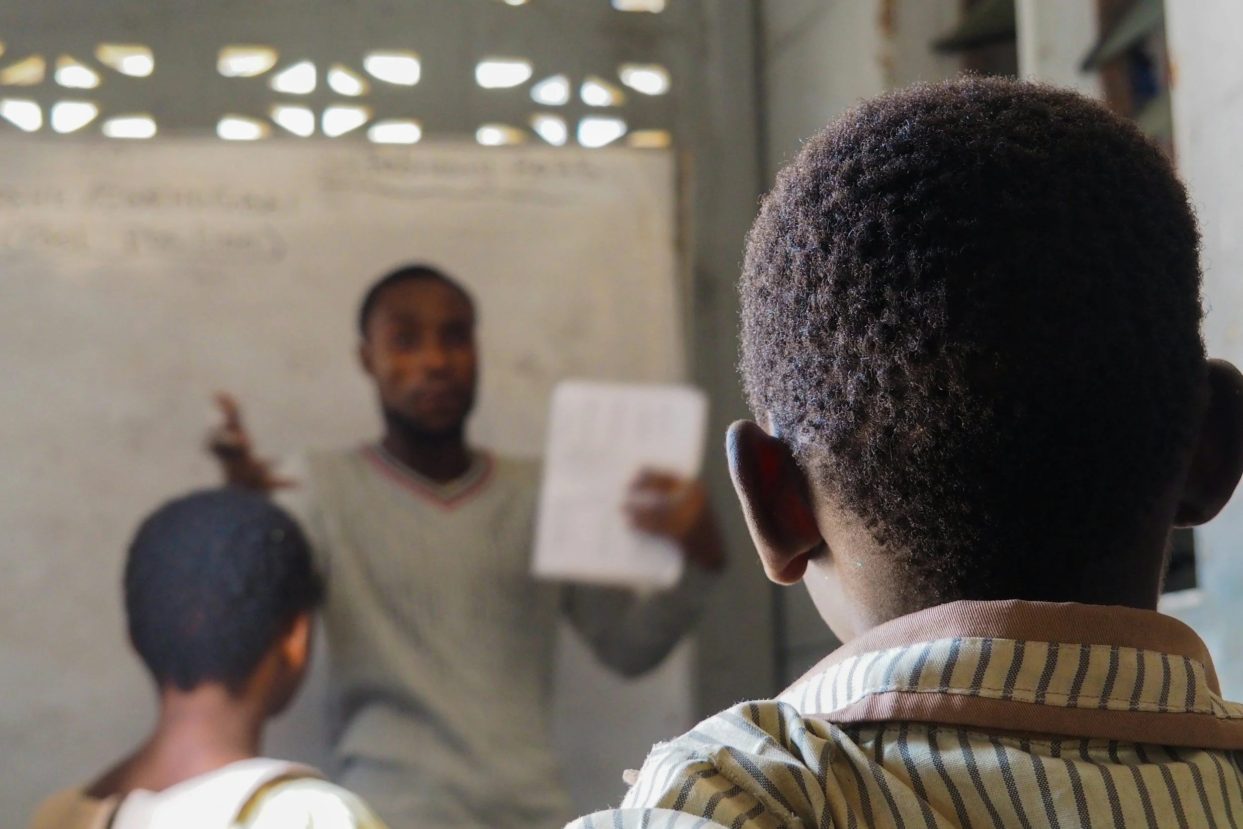 A child with curly hair and a striped shirt is looking at a man in the background, who is holding a document and gesturing with his hand in a classroom or meeting setting.
