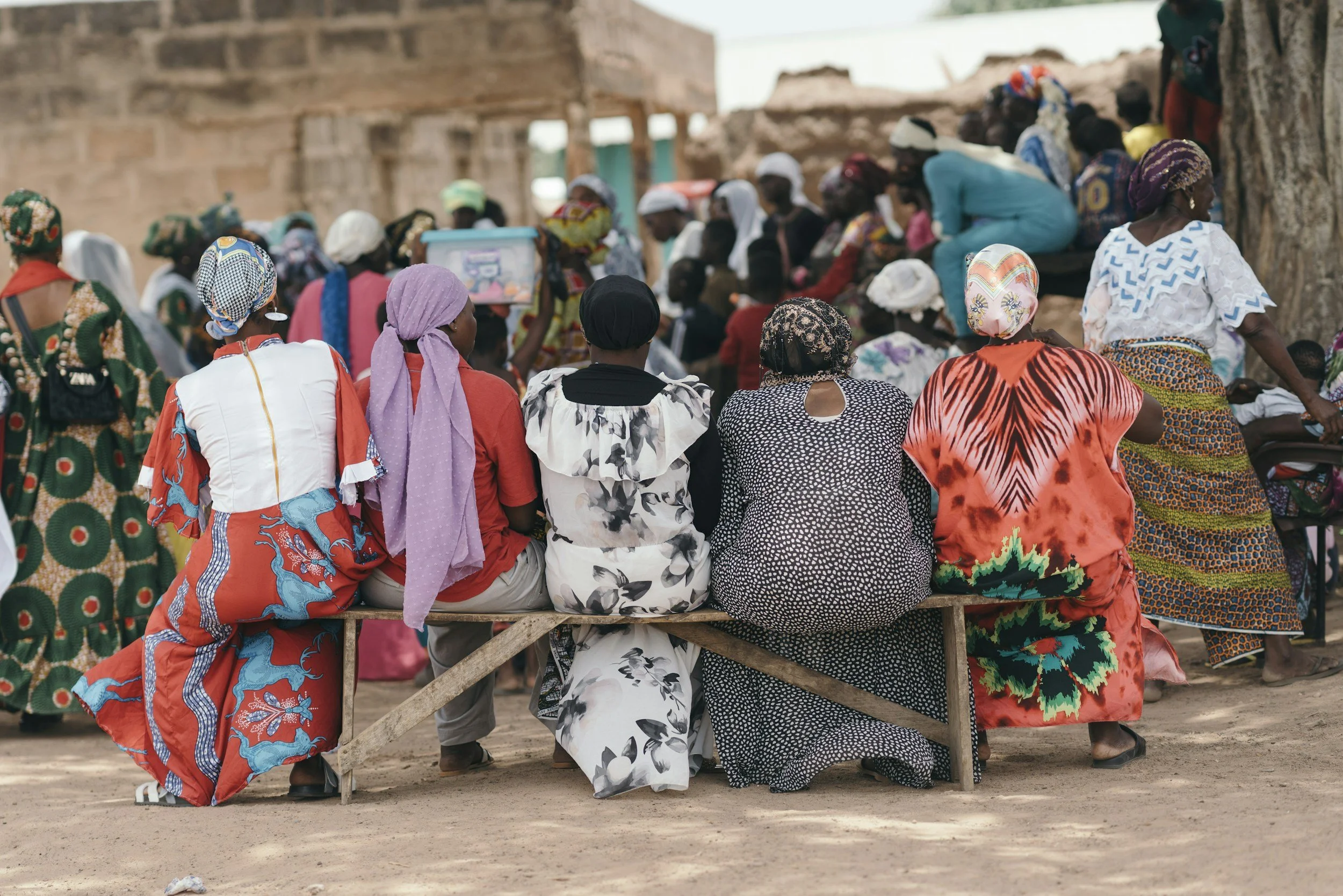 A group of people sitting on benches and gathered around in an outdoor setting with unfinished brick structures in the background. The people are dressed in colorful traditional clothing and head wraps, suggesting a community gathering or event.