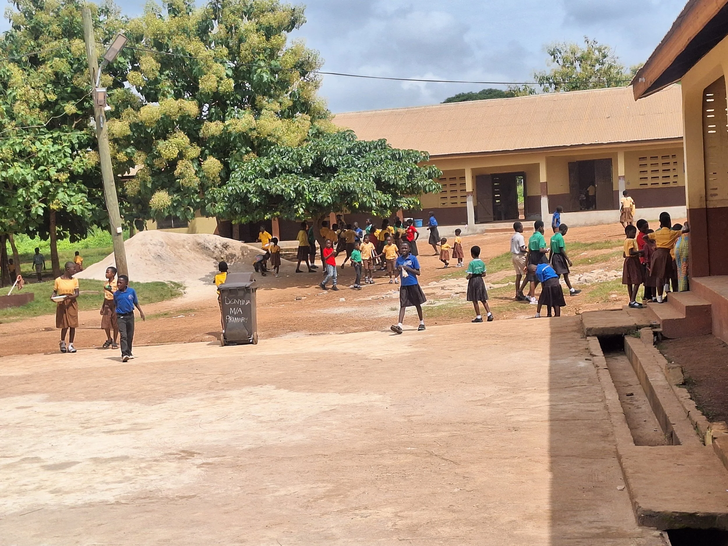 Schoolyard with students in uniforms, children playing, large trees, single-story school building, outdoor seating, and a trash bin with writing.