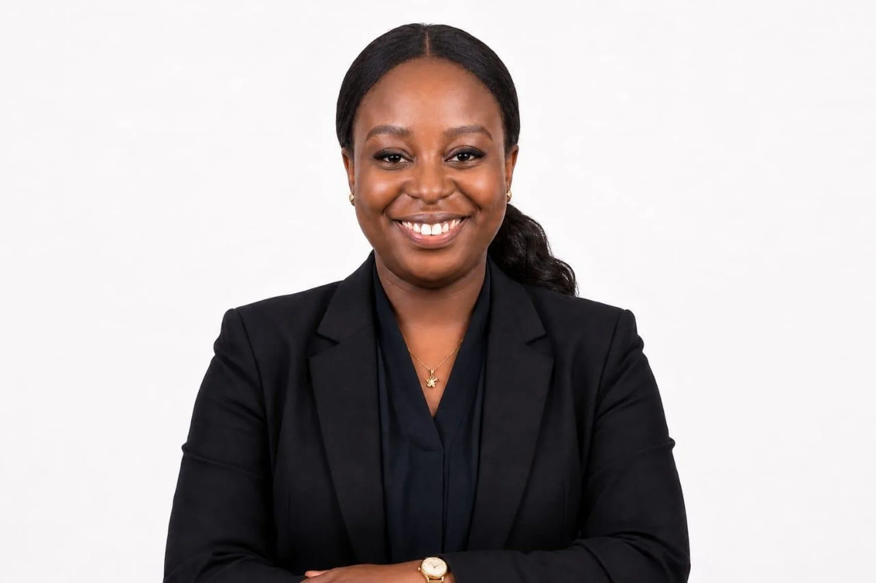 A smiling Black woman in a black blazer and navy blouse, with a gold necklace and watch, posing against a plain white background.
