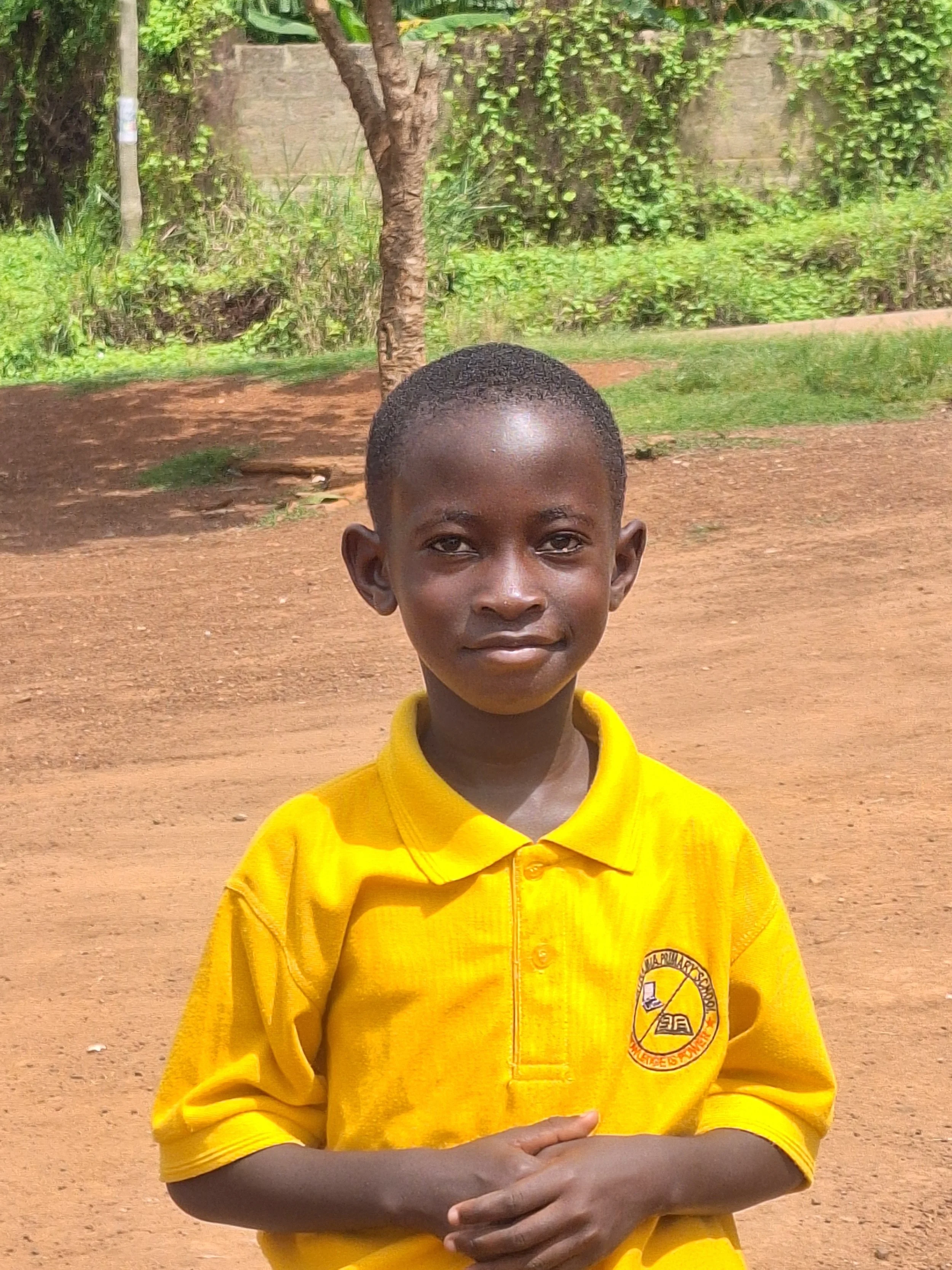 Portrait of a young boy with a short haircut, wearing a yellow shirt with a school emblem, standing outdoors on a dirt ground with trees and greenery in the background.