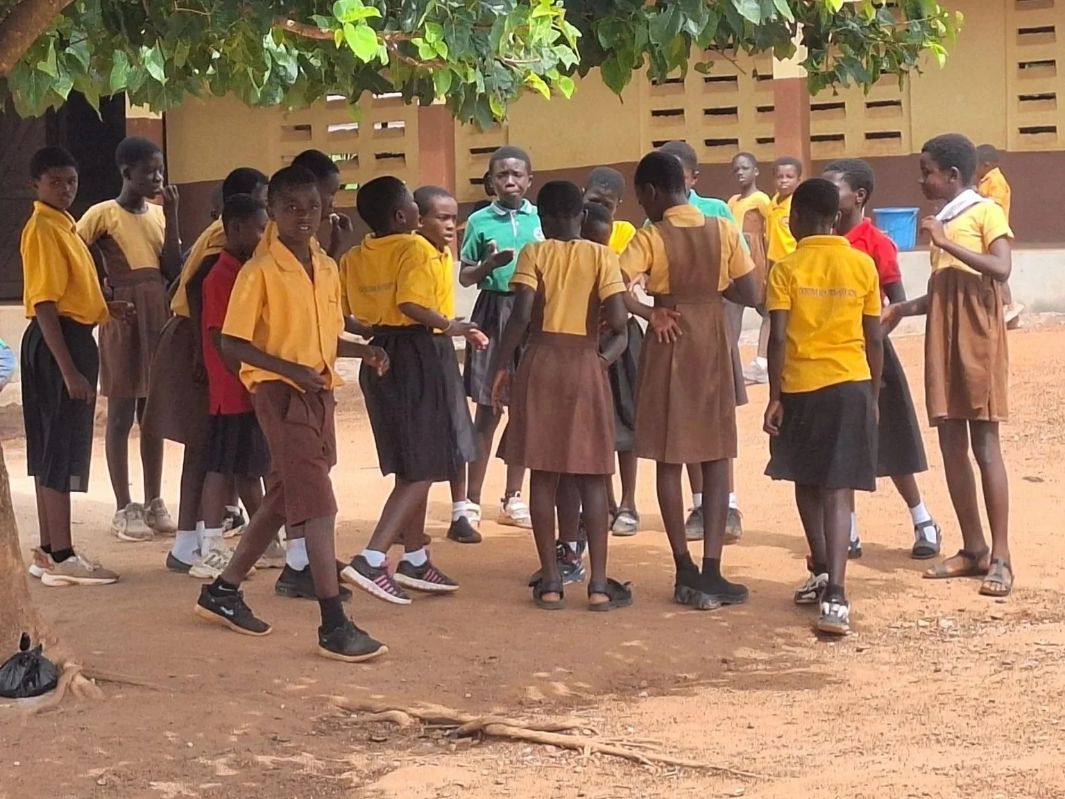 A group of schoolchildren wearing uniforms, some in yellow shirts and brown skirts, gathered outdoors under a tree, appearing to be engaged in a discussion or activity.