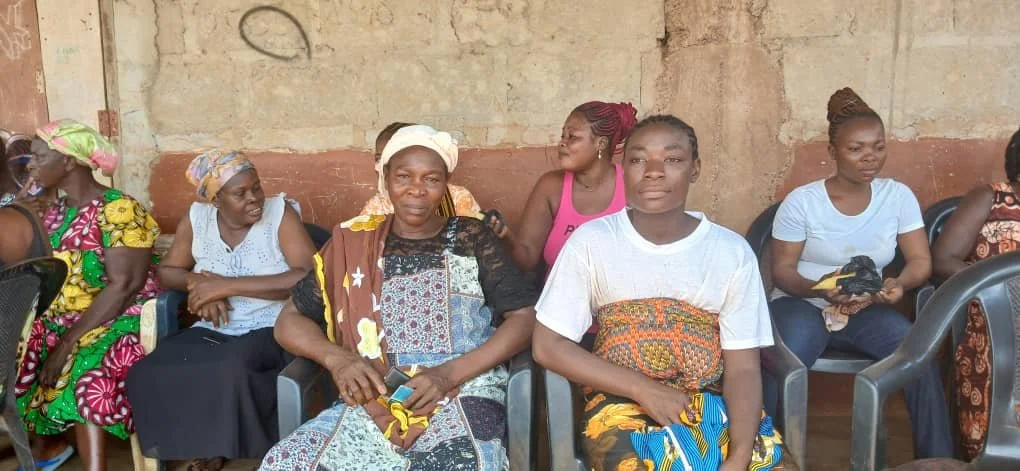 Group of women sitting on plastic chairs against a brick wall, some wearing colorful traditional African clothing and headscarves.