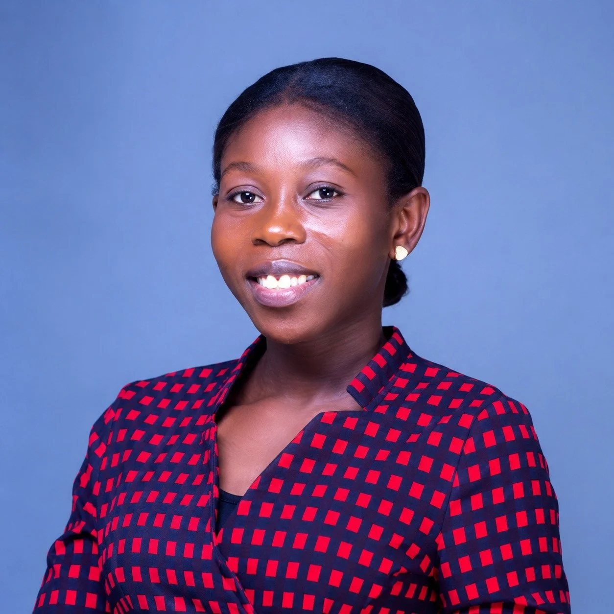 Portrait of a smiling African American woman with dark hair in a bun, wearing a red and black checkered blazer and gold earrings, against a blue background.
