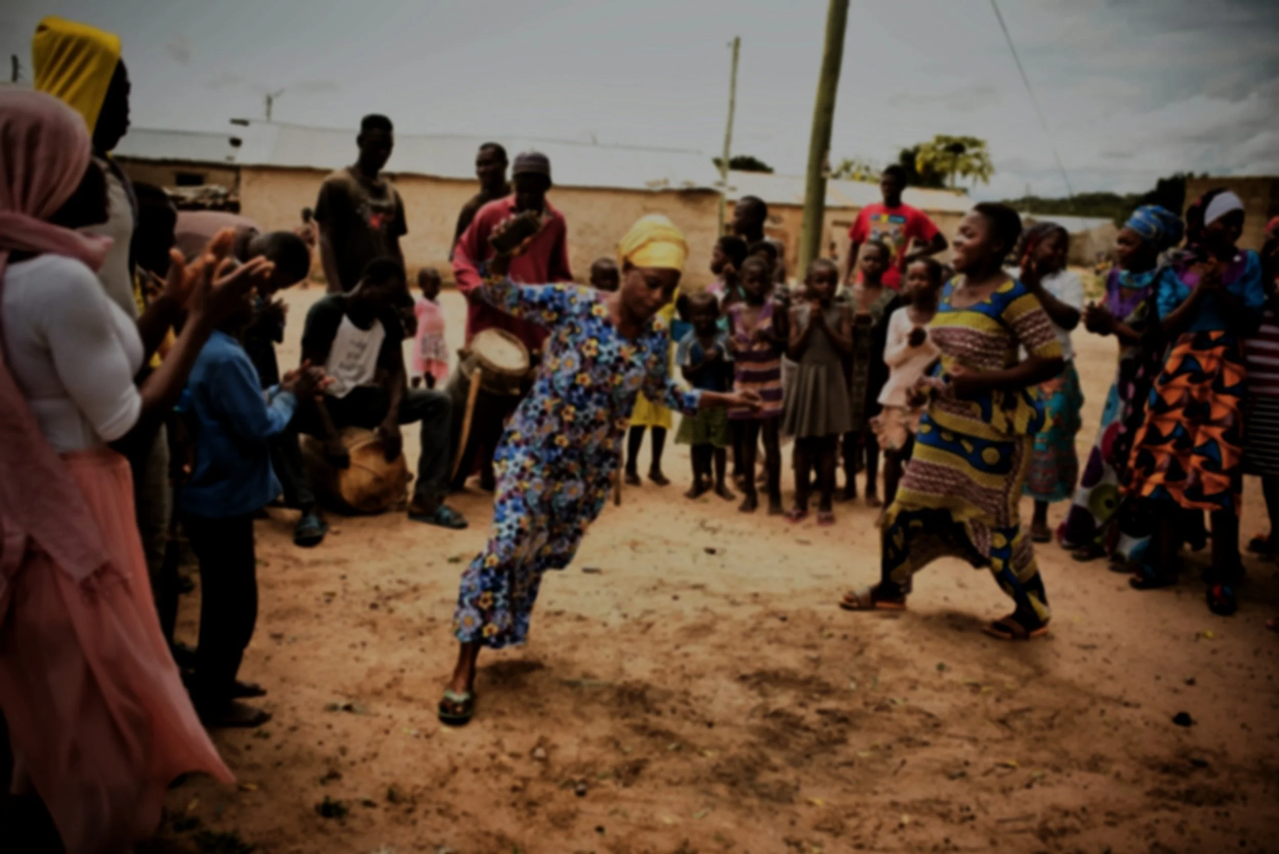 People gathered outdoors with some dancing and children watching. Women are dressed in colorful traditional clothing, and men are standing or sitting.