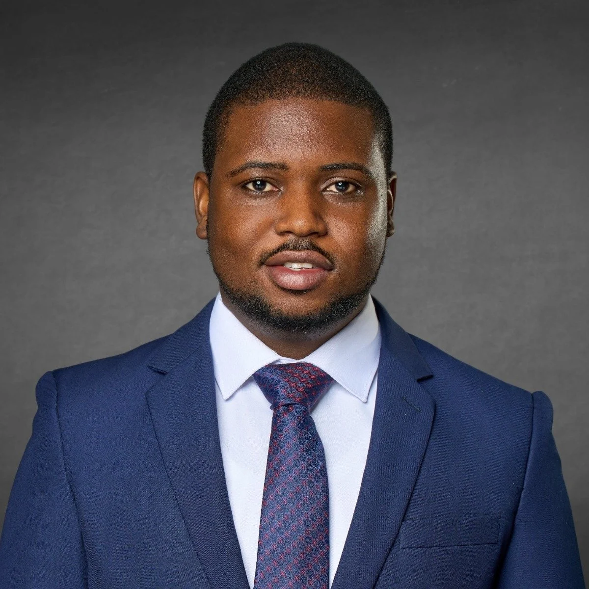 Professional portrait of a young African American man wearing a blue suit, white shirt, and patterned tie, against a gray background.