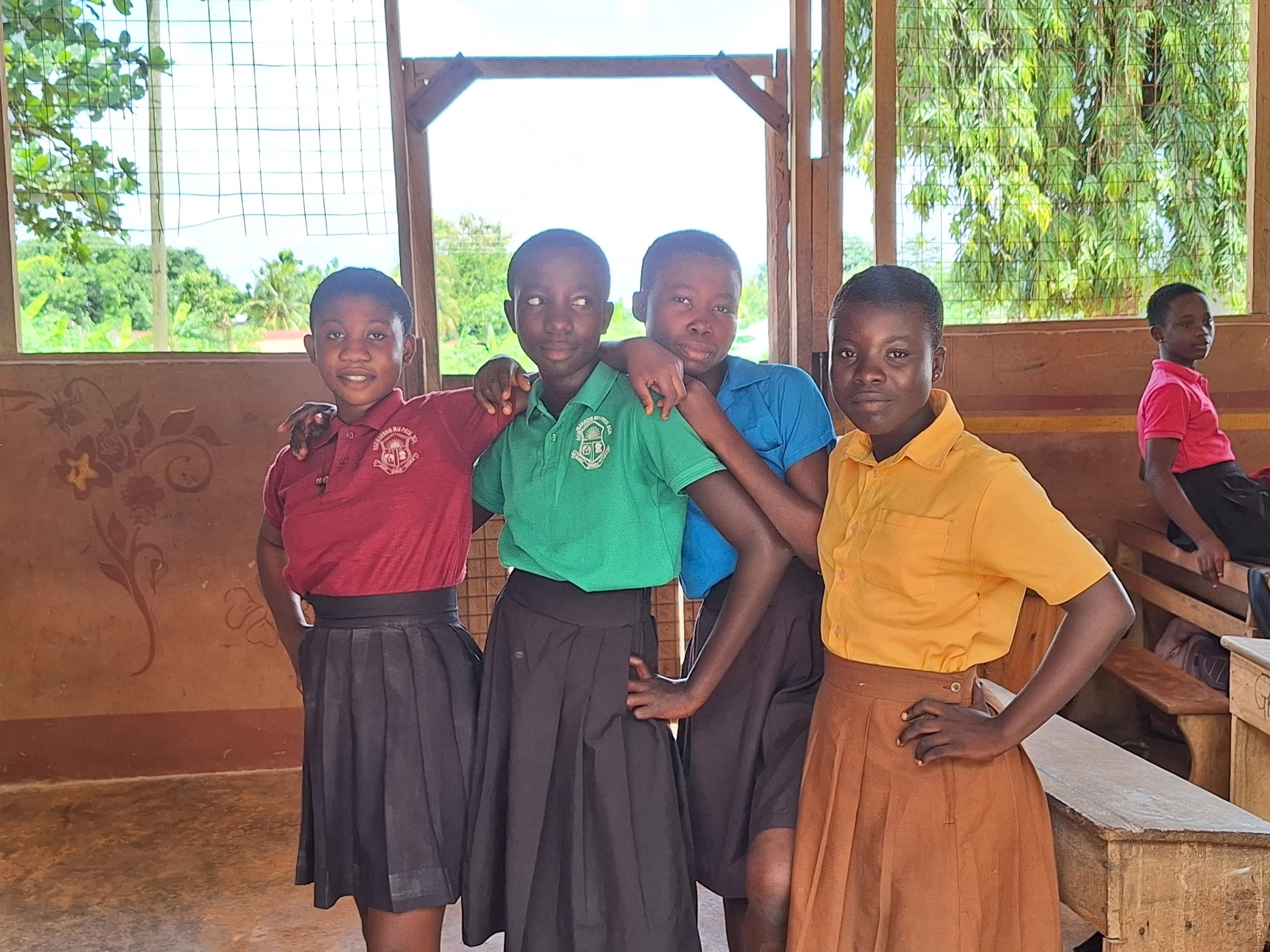 Group of five African children in school uniforms standing inside classroom, with a girl sitting on bench in background, greenery visible through windows.