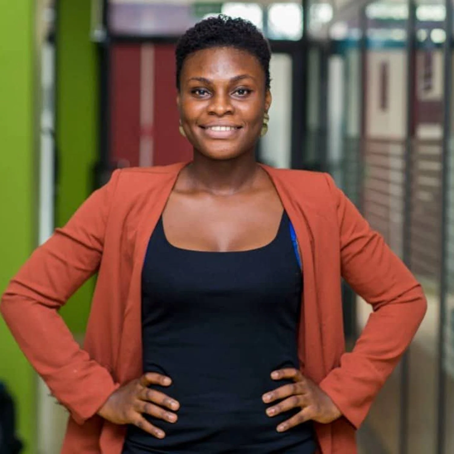 A confident woman standing in a modern office corridor with colorful walls, smiling at the camera with their hands on their hips.
