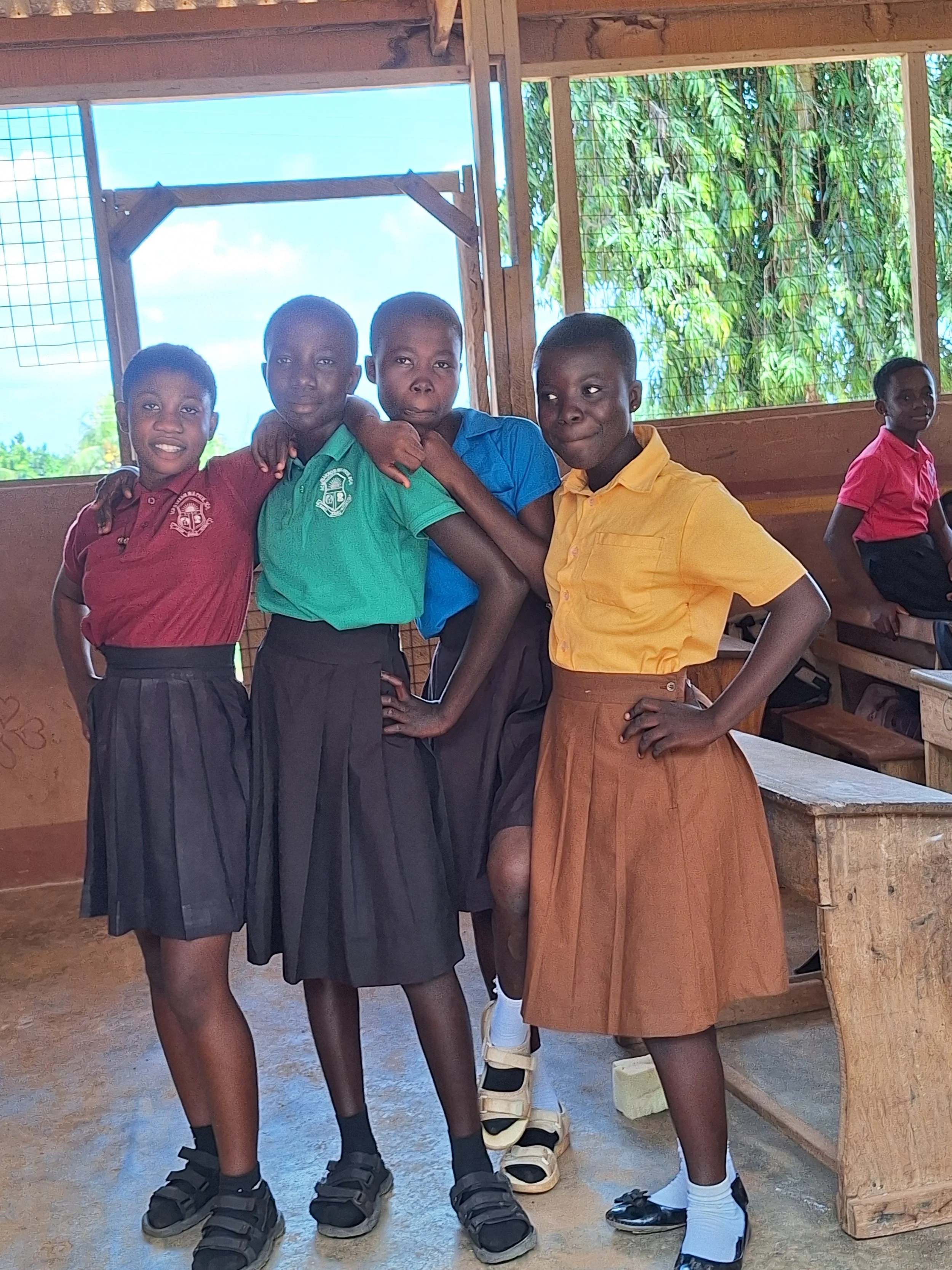 Four children standing inside a wooden classroom, four standing close and smiling, one child sitting on a bench in the background, with colorful uniforms and a sunny outdoor view visible through the open window.