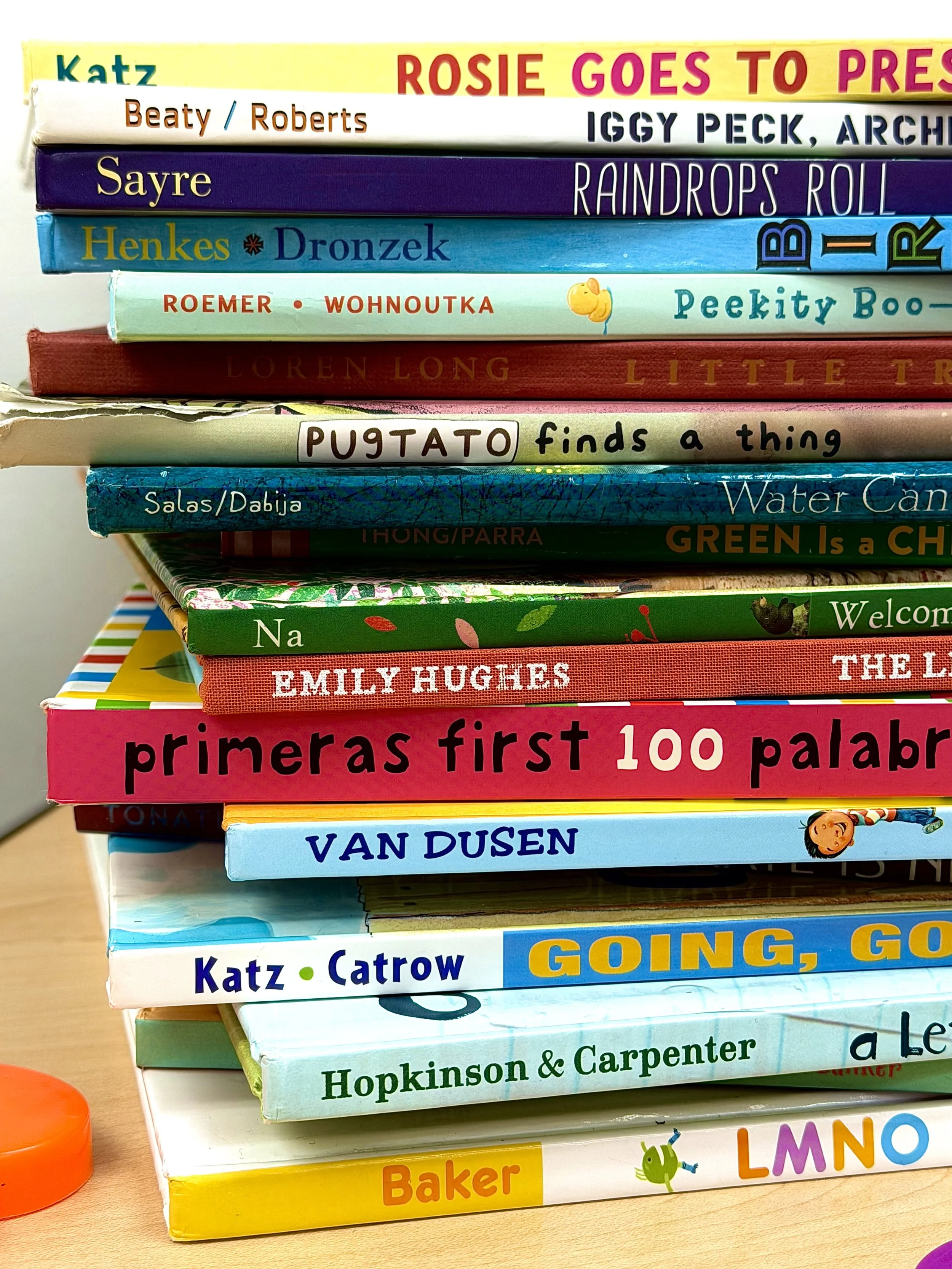 Stack of children's books with colorful covers including titles like 'Rosie Goes to Preschool', 'Iggy Peck, Architect', 'Raindrops Roll', 'Peekity Boo', 'Green Is a Chameleon', and 'First 100 Words'; placed on a wooden surface.