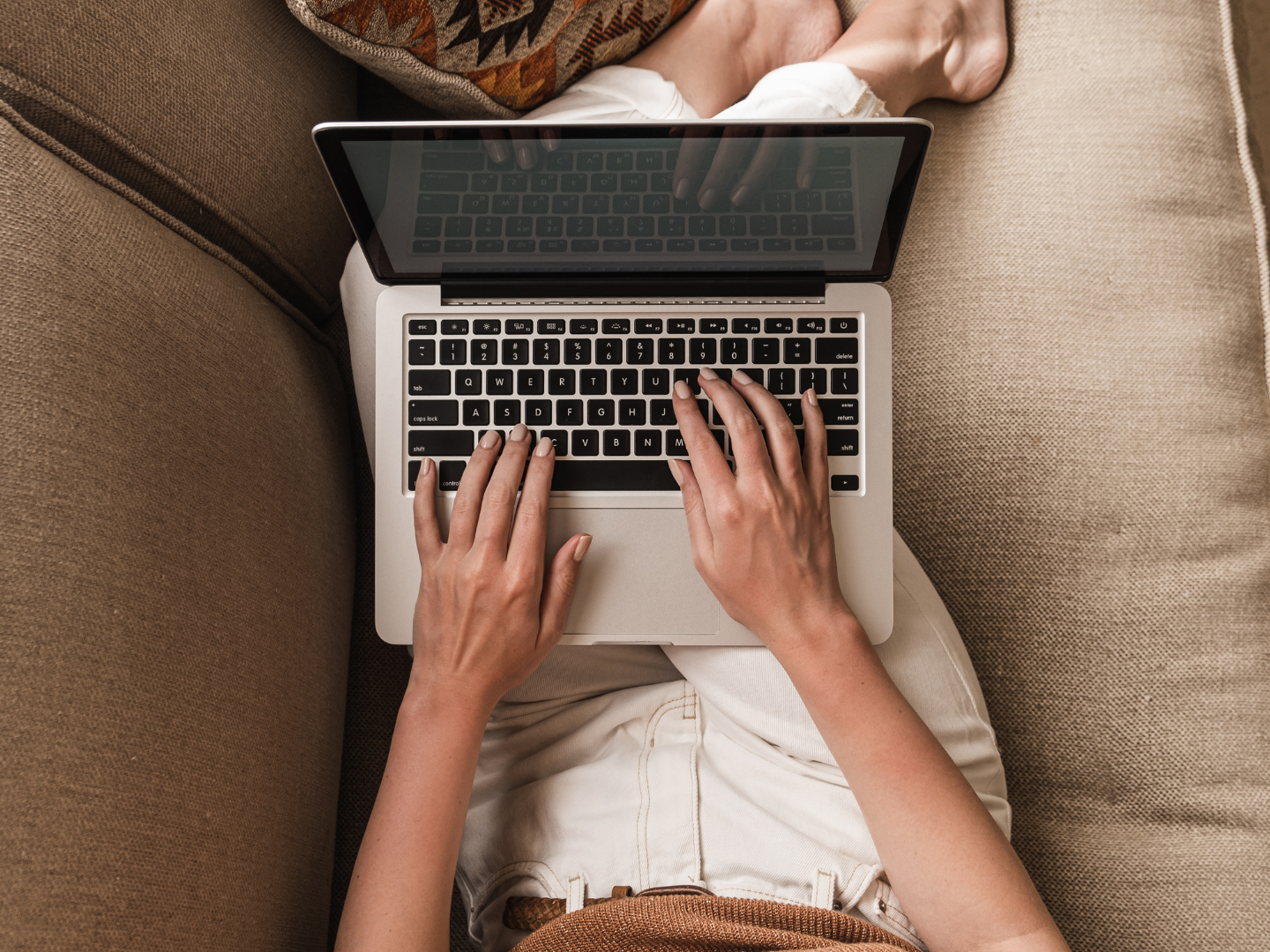 Person sitting on a beige sofa using a laptop, with feet tucked under a pillow, wearing a brown shirt and white pants.