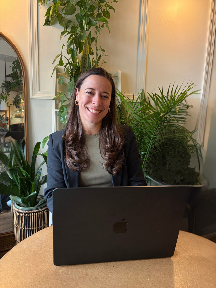 A smiling woman with long brown hair, wearing a black blazer and hoop earrings, sitting at a table with a black MacBook laptop open in front of her. She is indoors, surrounded by green houseplants, including a tall vine and a potted palm. There is a mirror and a bookcase with plants reflected in the background.