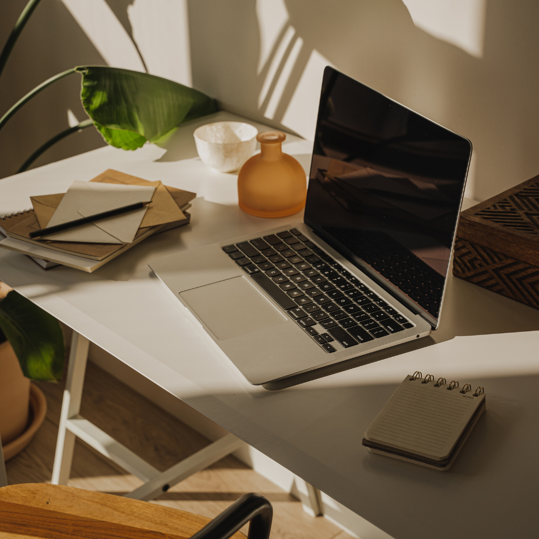 A white desk with an open laptop, a small notepad, a stack of books and envelopes, a pen, a decorative vase, a white bowl, and a patterned wooden box, illuminated by natural sunlight with shadows and green plant leaves nearby.