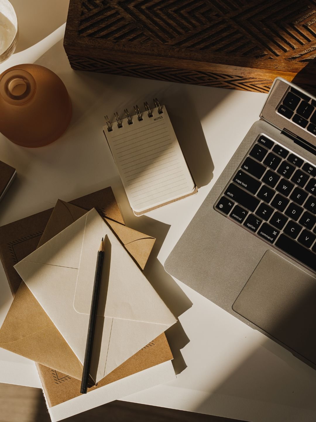 A work desk with a laptop, small notebook, envelopes, a pen, a decorative vase, and a wooden box, all on a white surface with sunlight casting shadows.
