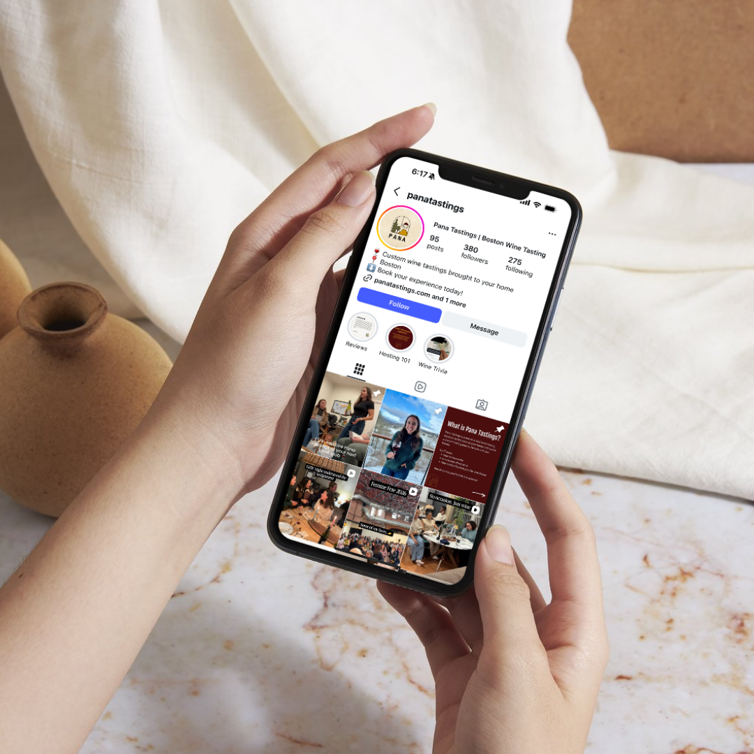 Person holding a smartphone displaying the Instagram profile of Pana Tastings with images of wine tastings and events, on a marble table next to two pottery vases.