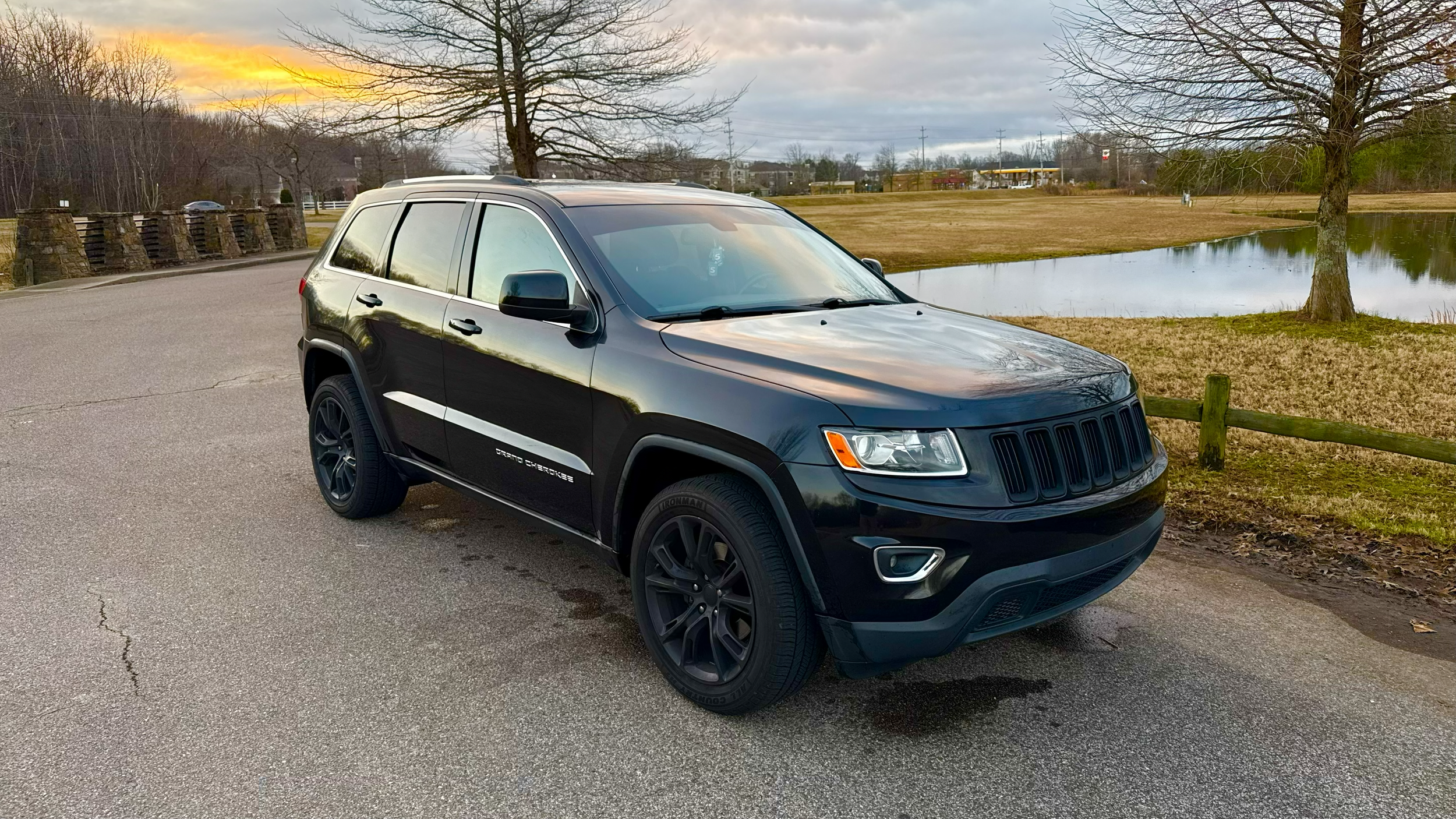 Black Jeep Grand Cherokee parked on a paved road next to a grassy area with a pond, trees, and a sunset sky in the background.