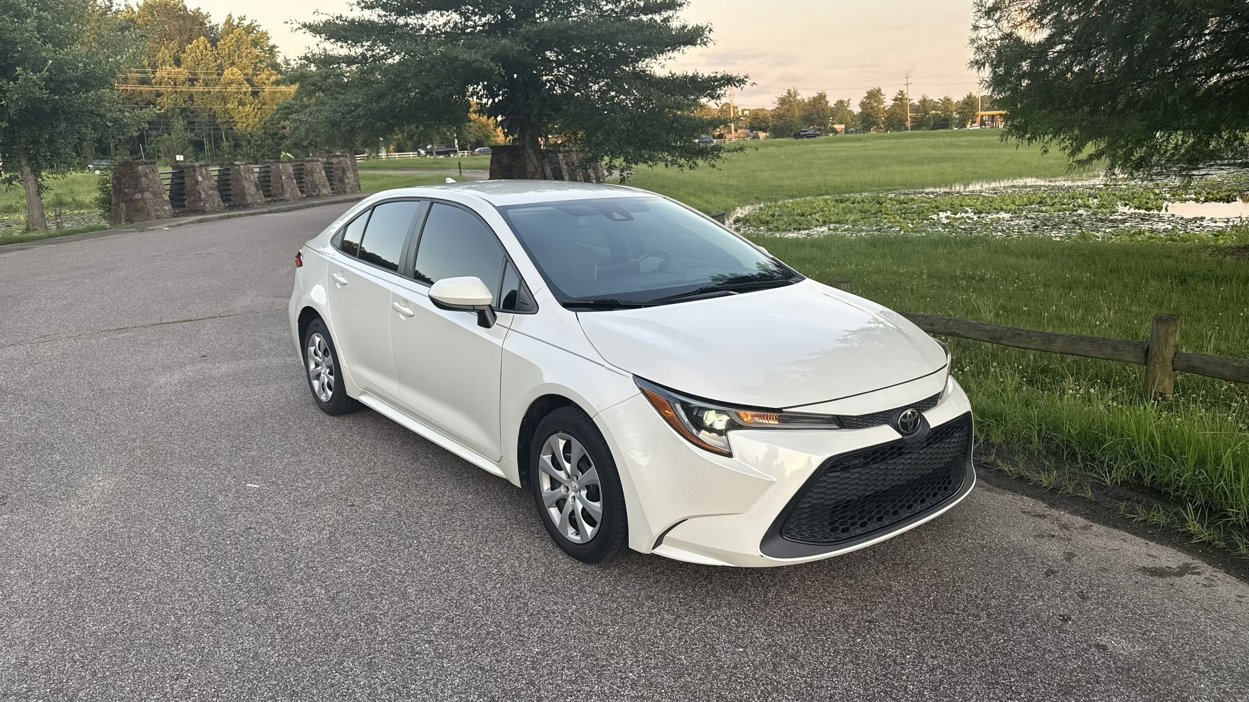 A white Toyota sedan parked on a gravel area near a grassy park with trees and a pond in the background during late afternoon or early evening.
