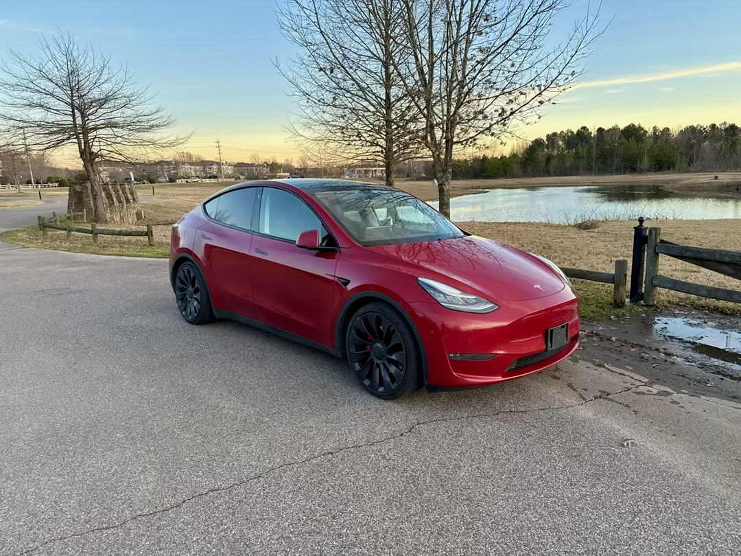 A red Tesla Model Y parked on the side of a paved road near a small lake, with leafless trees and a sunset sky in the background.