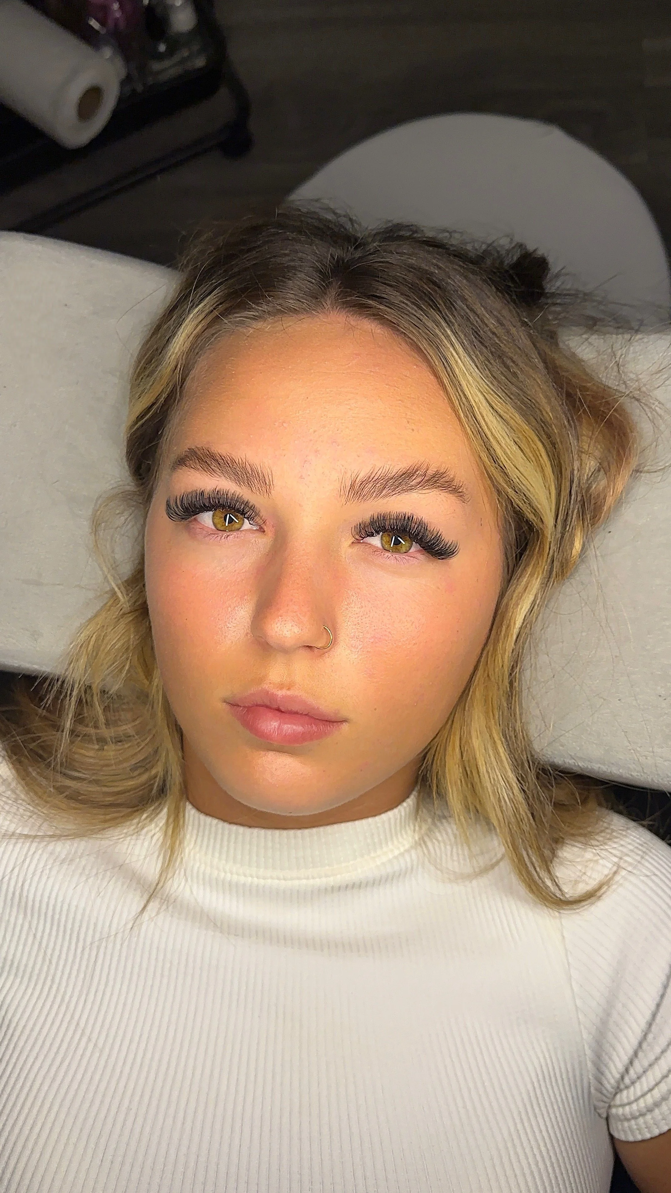 Close-up of a young woman with blonde hair, wearing a white top, lying on a table with her head on a pillow, with eyelash extensions, a gold nose ring, and makeup.
