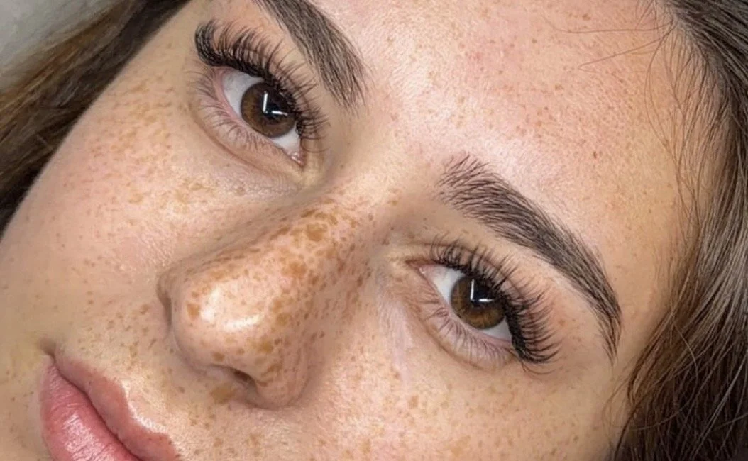 Close-up of a woman's face, showing her brown eyes, long eyelashes, eyebrows, and freckled skin.