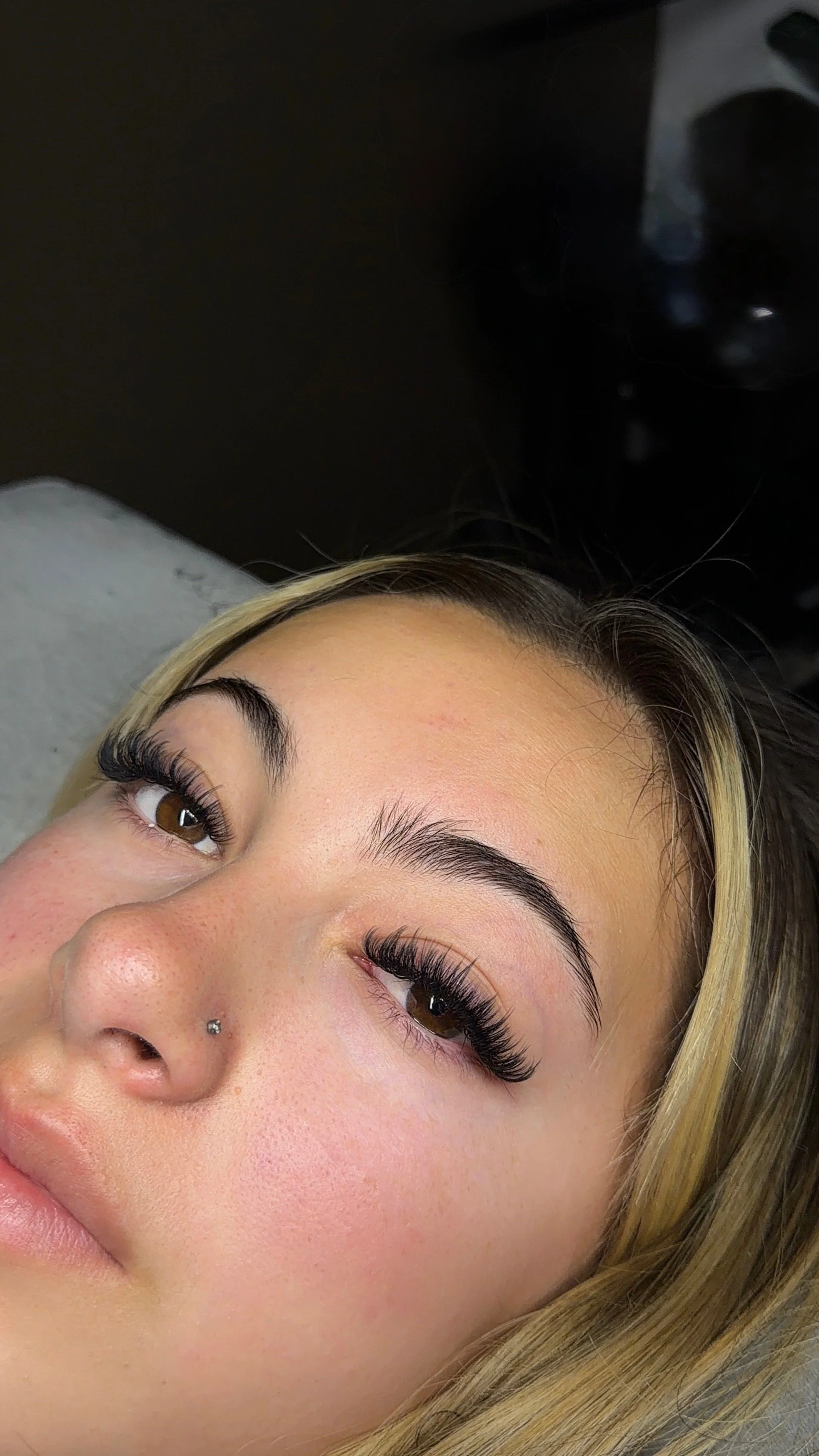 Close-up of a woman's face with brown eyes, long eyelashes, blonde hair, and a nose piercing, lying on a gray surface.