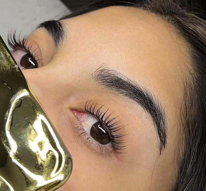 Close-up of a woman's face showing her eye, eyebrow, and part of her nose, with her holding a gold-colored reflective object near her face.