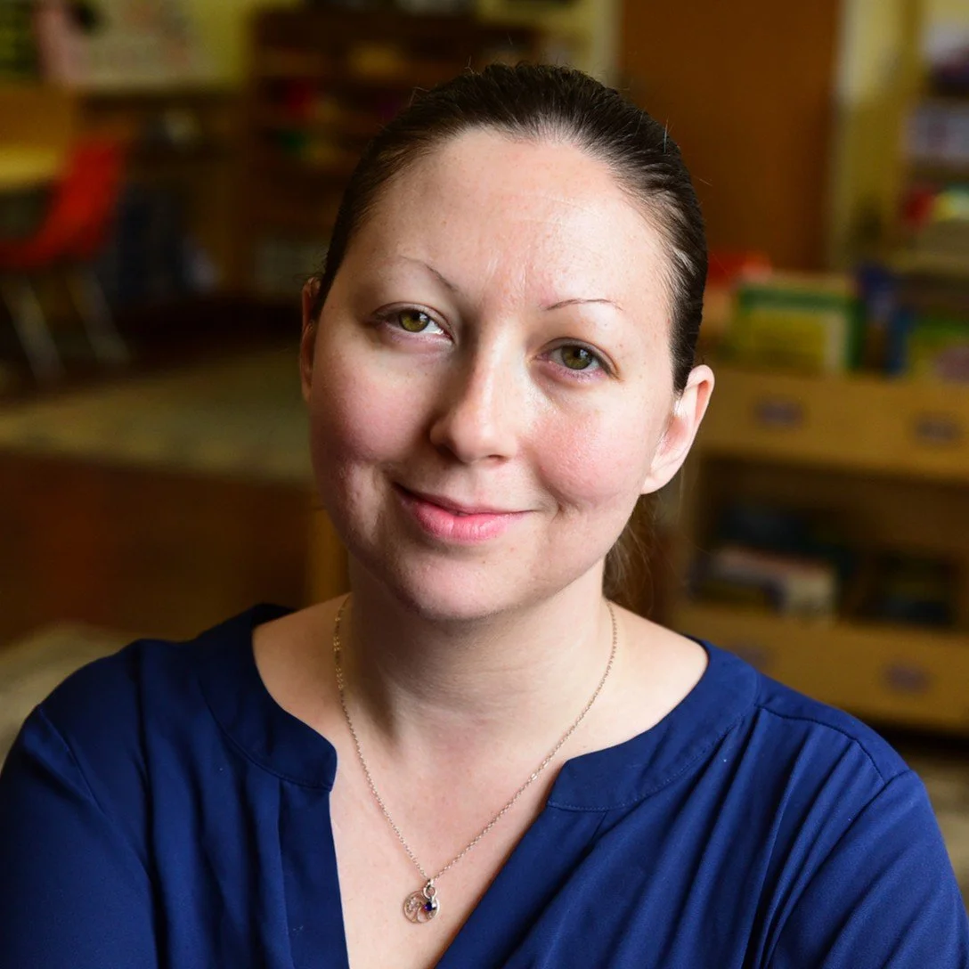A woman with dark brown hair pulled back, wearing a navy blue top and a delicate necklace, smiling softly in an indoor setting with blurred bookshelves and colorful books in the background.