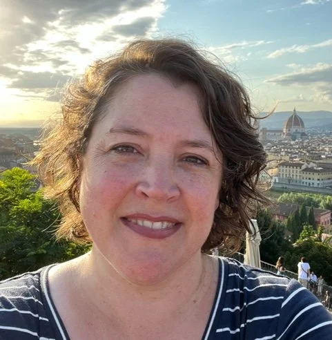 A woman taking a selfie outdoors with the Florence skyline, including the Florence Cathedral, in the background during sunset.