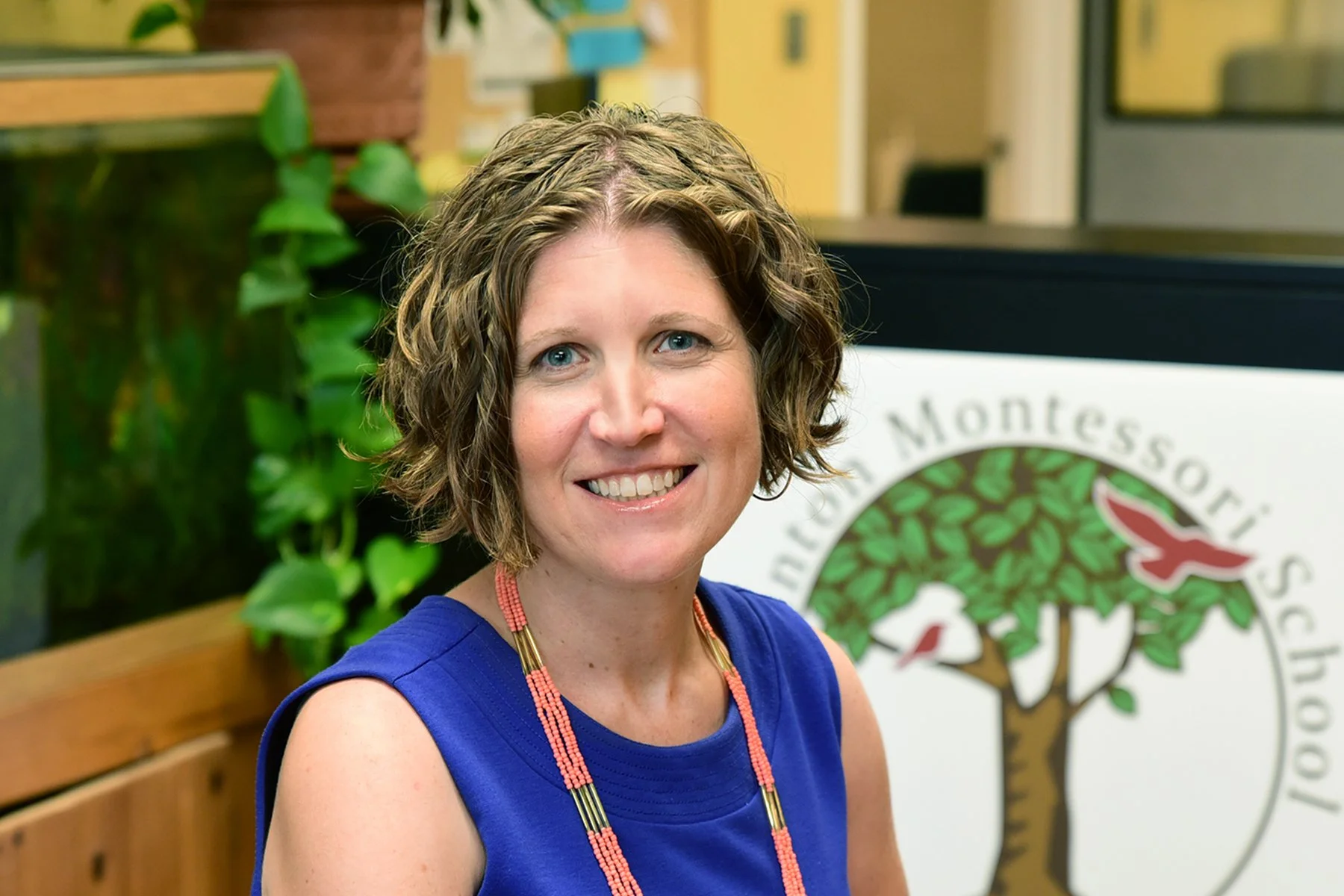 A woman with short curly light brown hair and blue eyes smiling, wearing a royal blue dress and a beaded necklace, sitting in front of a sign that reads 'Montesori' with a leafy tree logo, in an indoor setting with a green plant and wooden bench in the background.