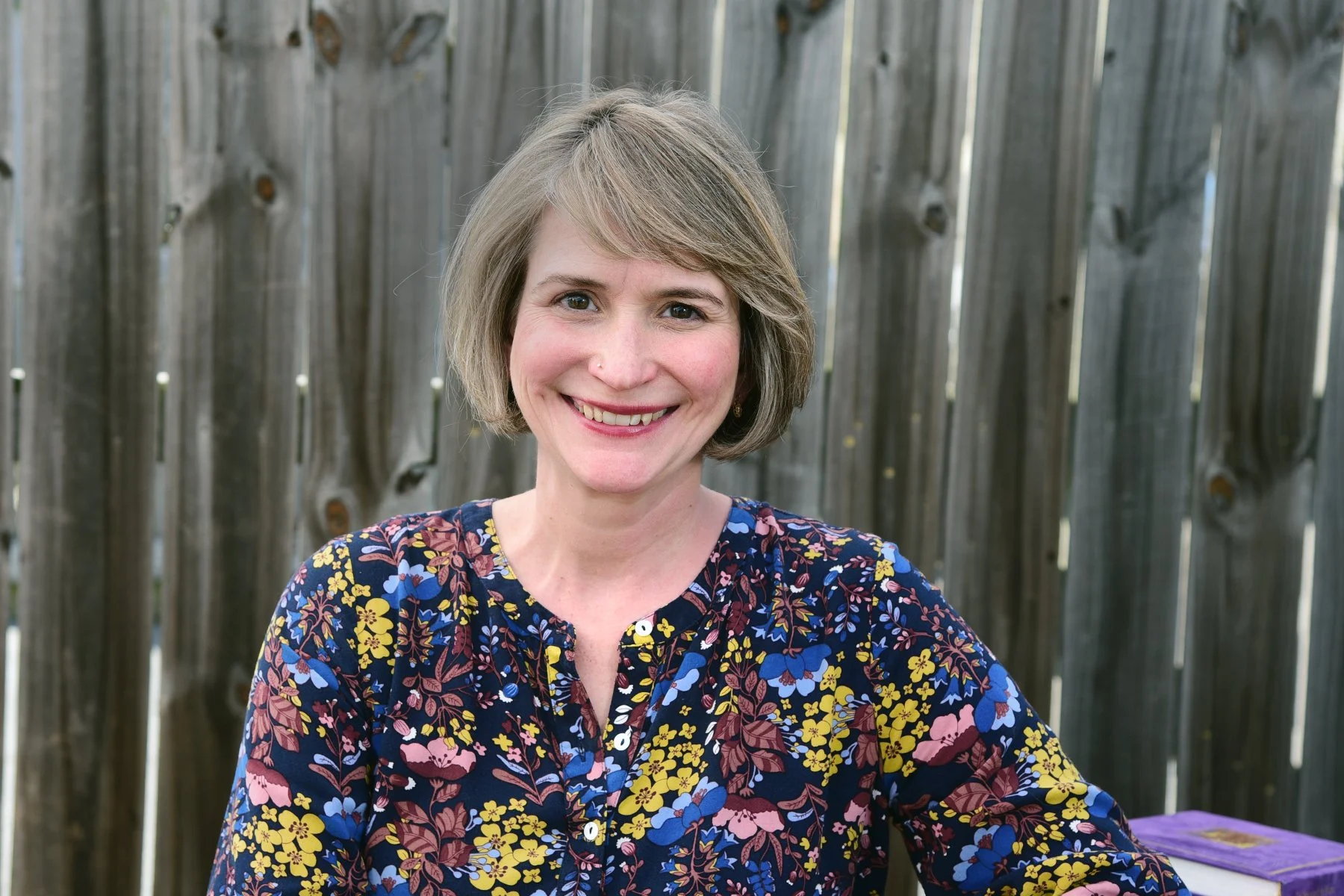A woman with short, light brown hair smiling, wearing a colorful floral shirt, sitting outdoors with a wooden fence in the background.