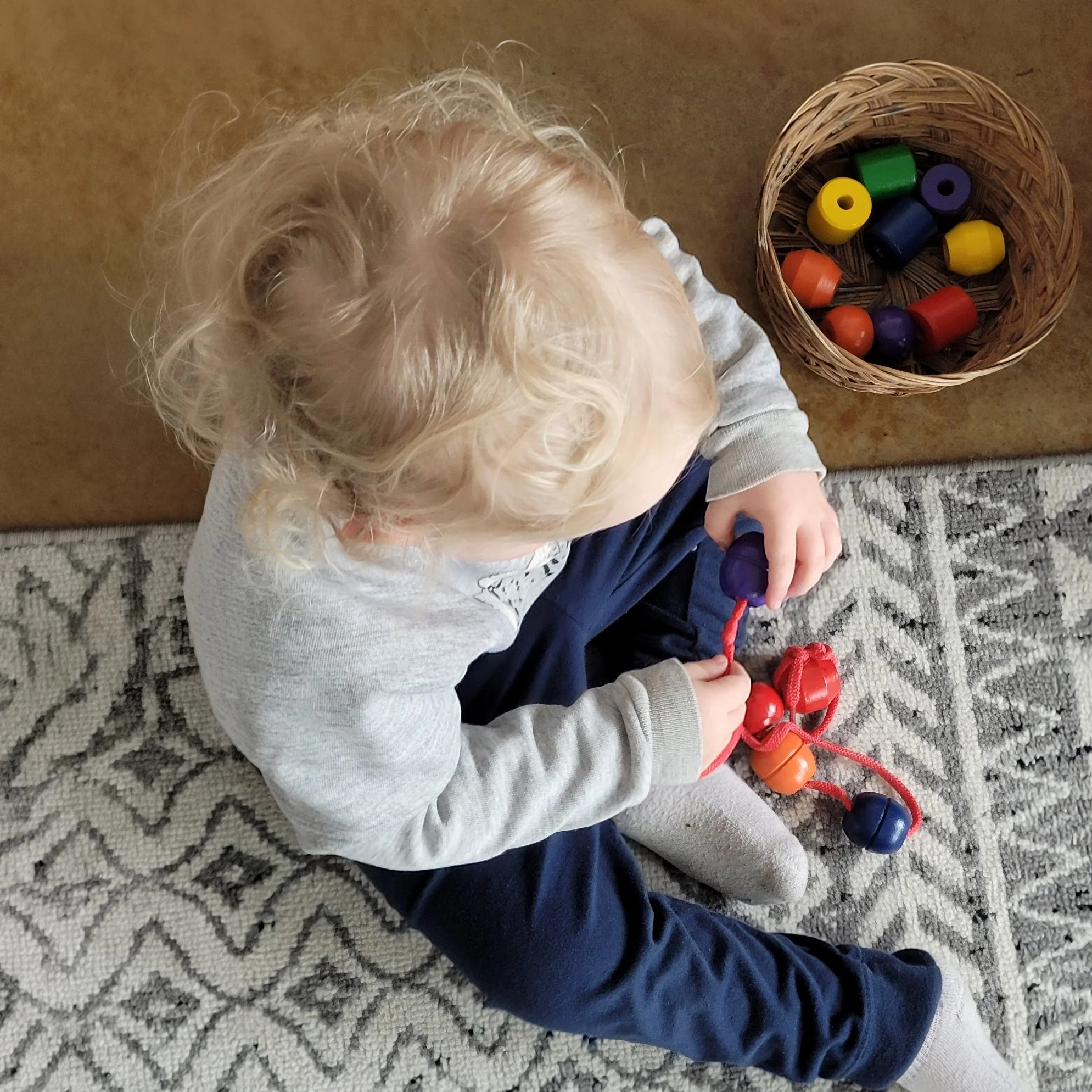 Top view of a young child with curly blonde hair sitting on a textured gray carpet, holding a red toy. To the child's right is a round wicker basket filled with colorful wooden toy bottles in yellow, green, purple, blue, orange, and red.