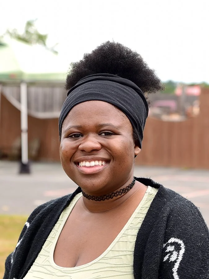 A young woman with dark skin and natural hair styled in an afro, wearing a black headband, a black choker necklace, a light-colored top, and a black cardigan, smiling outdoors with a blurred fence and trees in the background.