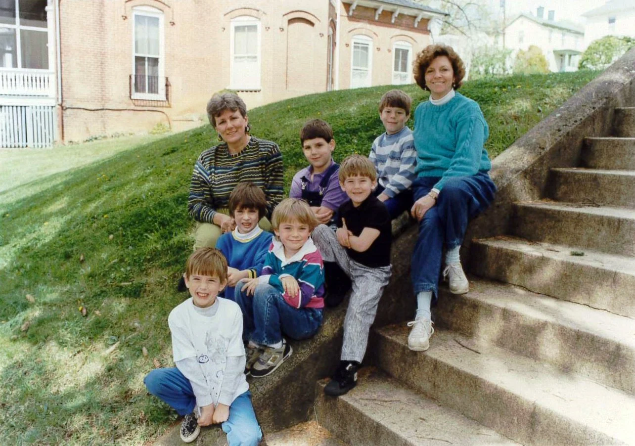 A group of children and two women sitting on steps outdoors, in front of grass and brick buildings, during daytime.