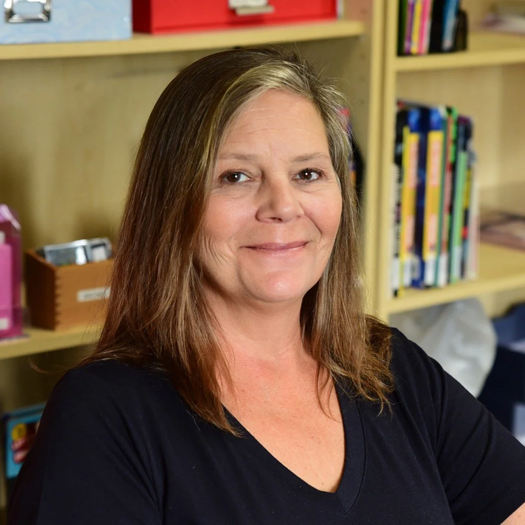 A woman with shoulder-length brown hair and a black shirt standing in a room with yellow shelves filled with colorful books.