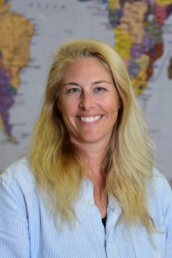 A woman with long blonde hair smiling in front of a world map.