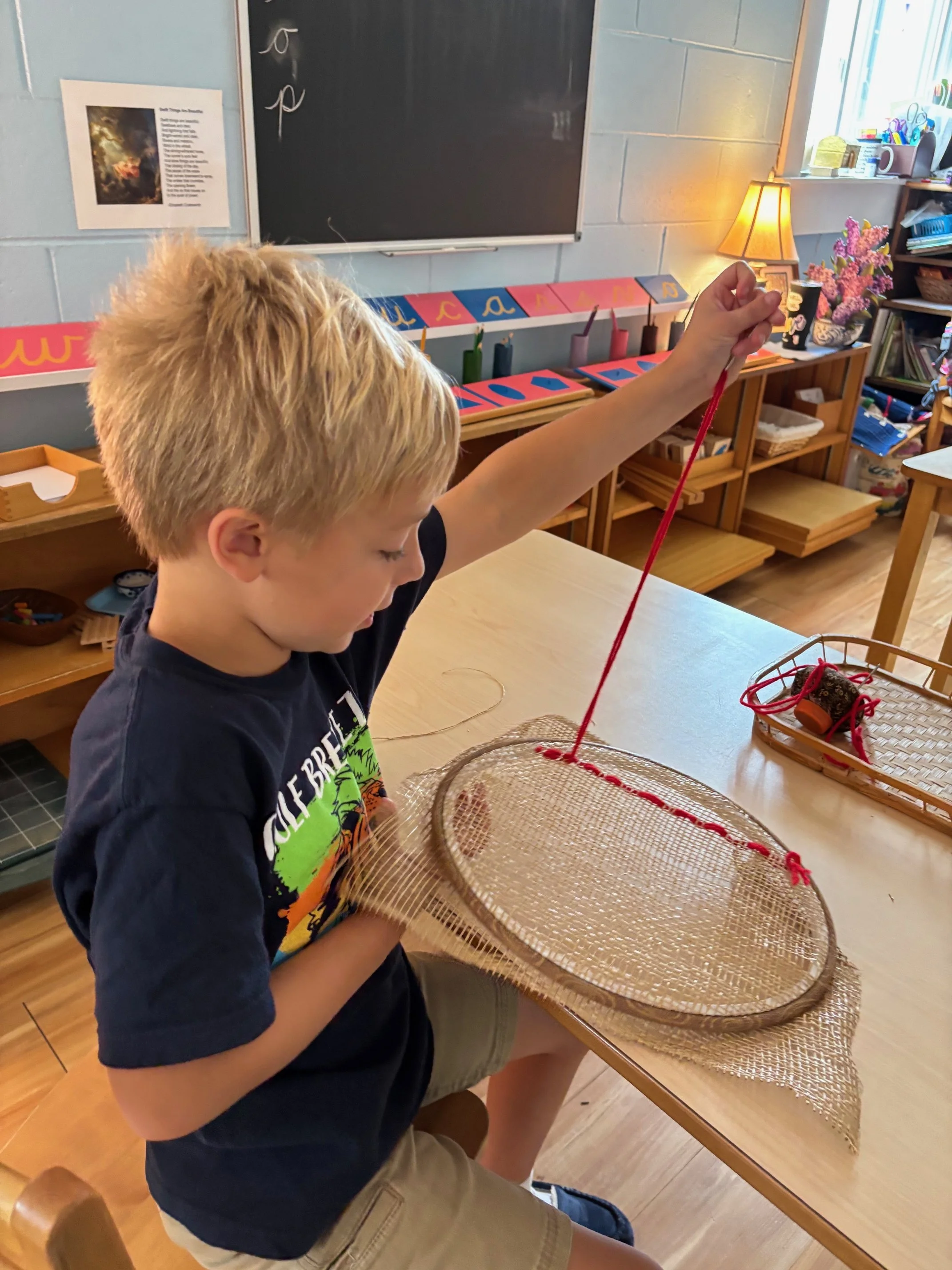 A young boy with blonde hair wearing a black T-shirt and beige shorts, sitting at a table in a classroom, holding a circular woven basket with a red string inside. He appears to be engaged in a craft activity involving the basket.