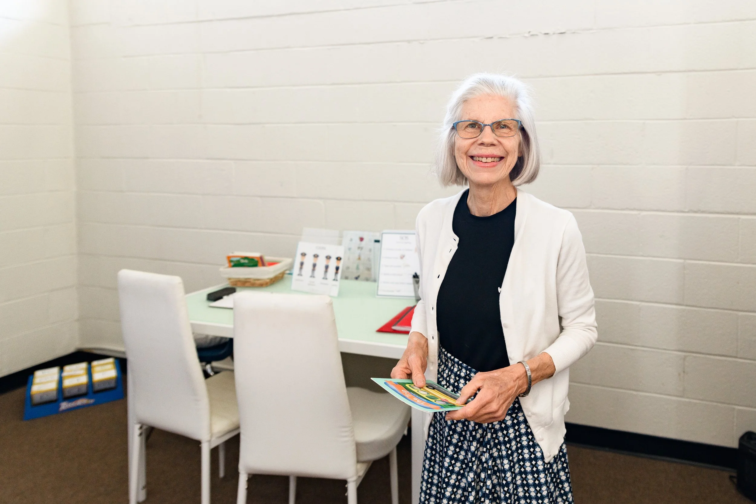 Smiling elderly woman with gray hair and glasses holding a brochure inside a room with white walls and chairs.