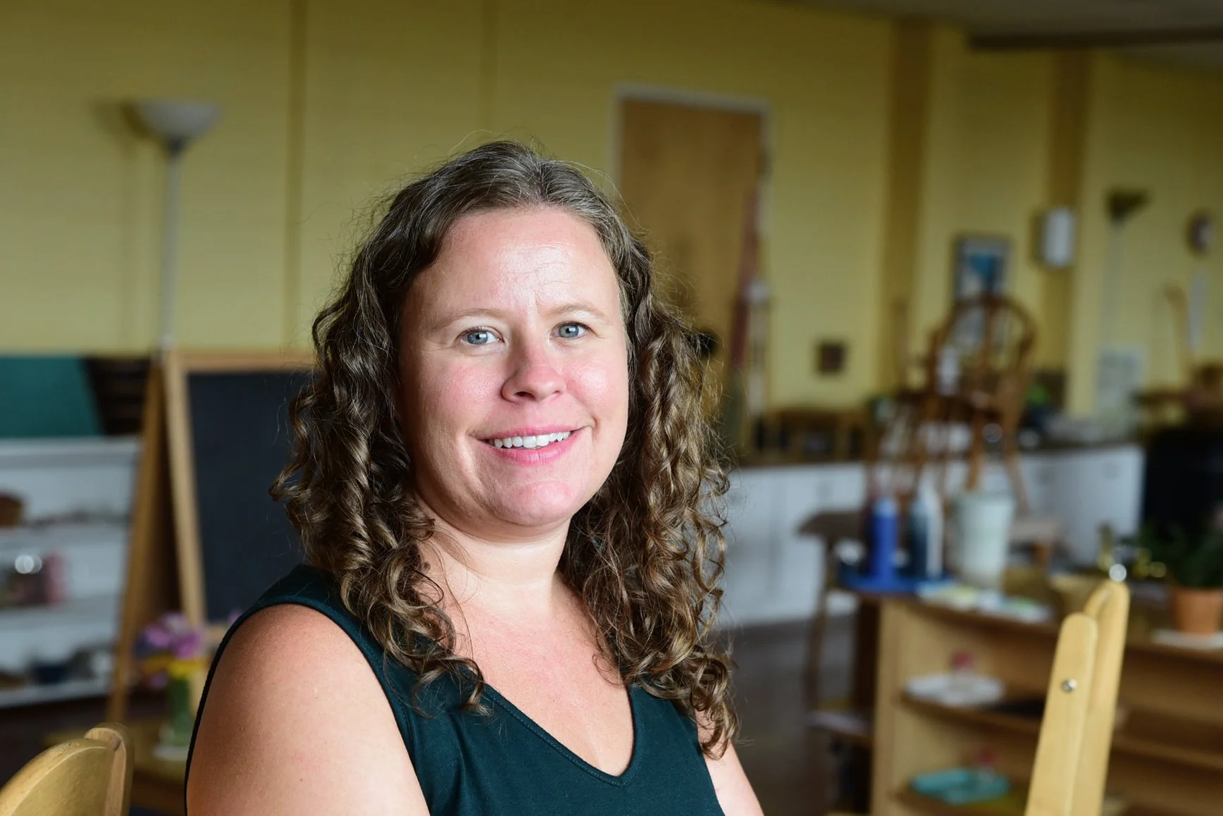 A woman with curly brown hair and blue eyes smiling at the camera in a room with yellow walls and wooden furniture, including chairs, shelves, and a table.