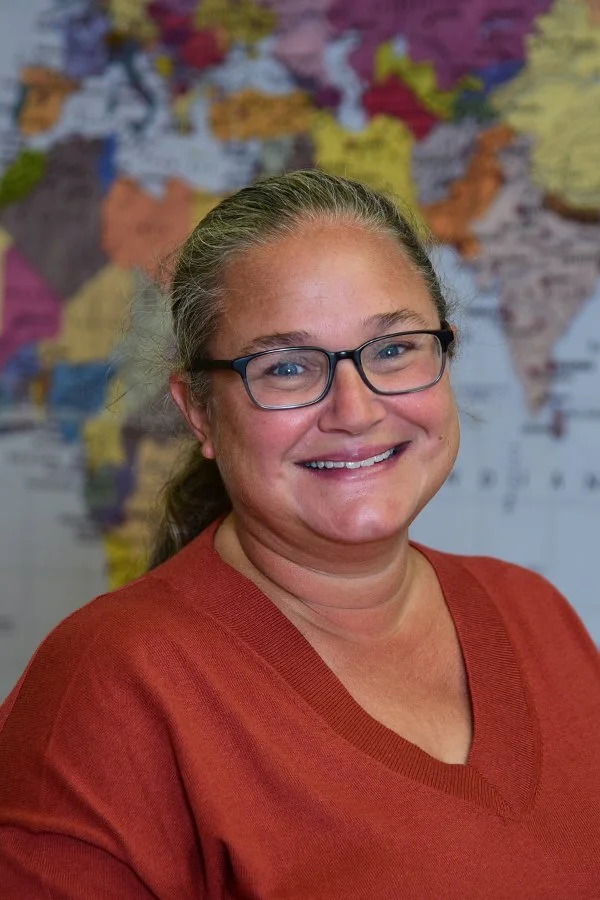 A smiling woman with glasses wearing a red shirt standing in front of a colorful world map.