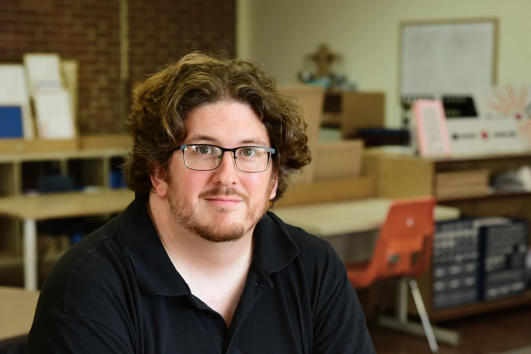 A man with curly brown hair, glasses, and a beard, wearing a black shirt, sitting in a classroom or office environment with shelves, boxes, and a whiteboard in the background.